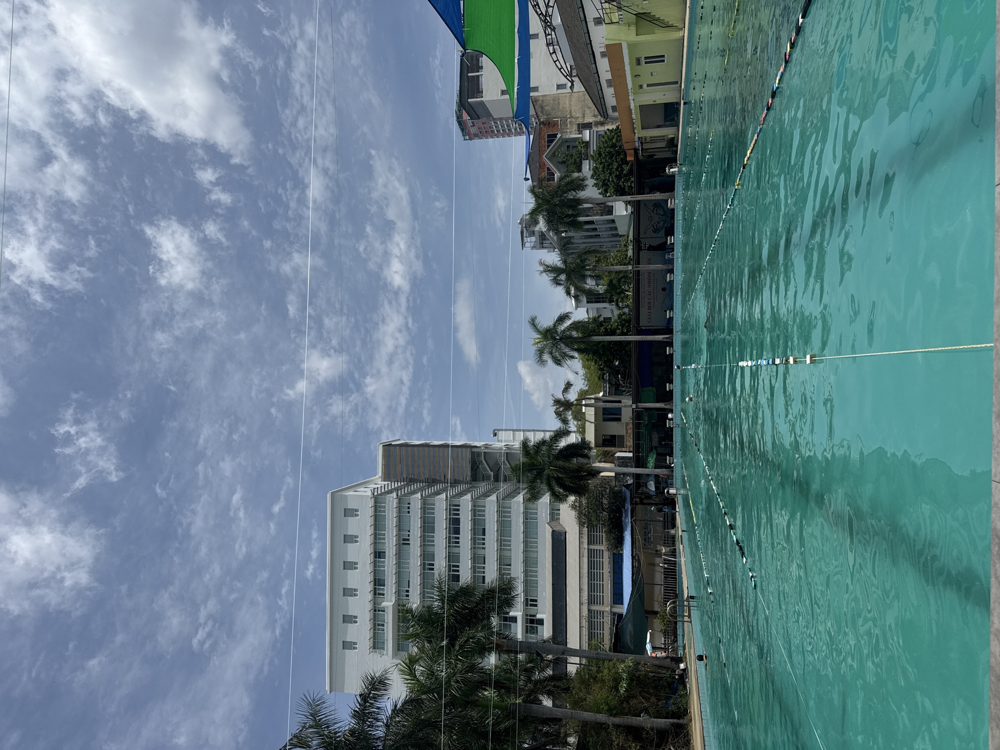 The image shows an outdoor swimming pool in Nha Trang Ward, Vietnam. The pool is a bright turquoise color and divided into lanes by ropes with buoys. There are several swimmers in the pool, some actively swimming and others appearing to be resting. Palm trees are visible on the left side of the image, and a large, modern building stands behind them. Other buildings, including a brightly colored green and yellow structure, are visible on the right side of the image. The sky is cloudy with patches of blue. The overall scene depicts a public swimming facility on a cloudy day.