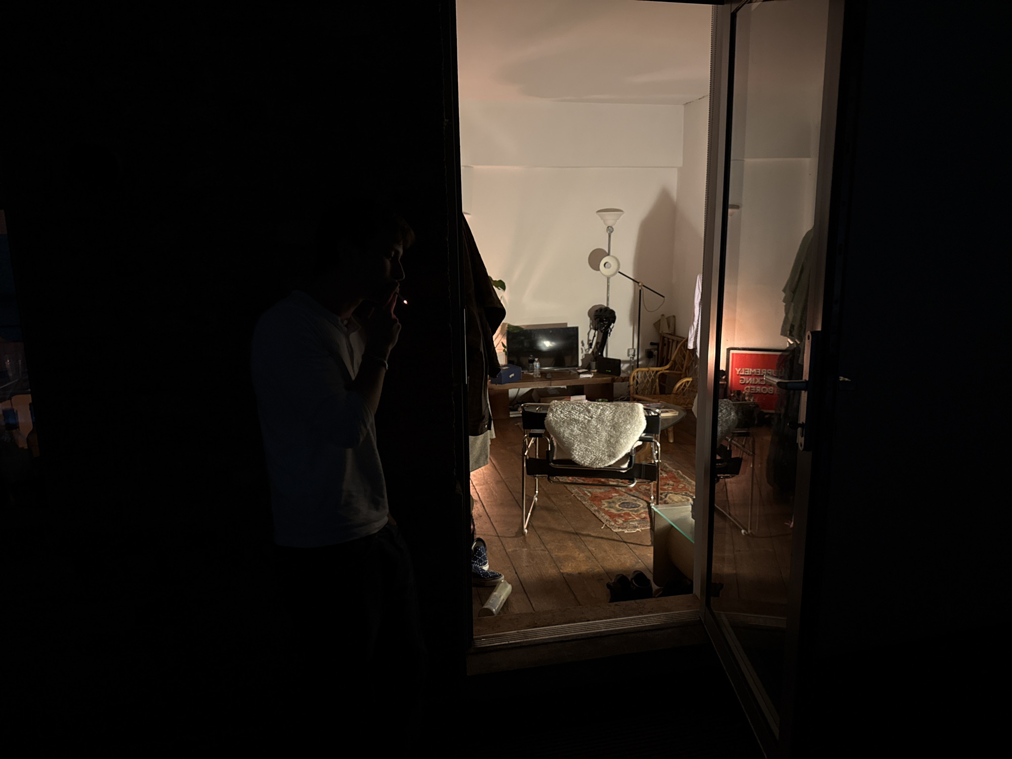 A dimly lit interior is seen through an open doorway. A man stands in the darkened foreground, appearing to smoke, partially obscured by the door frame. The illuminated room beyond features a chair with a white cushion, a television on a wooden stand, a floor lamp, and various other pieces of furniture, including what appears to be a rattan chair. A poster with reverse text is visible. The room is bathed in warm artificial light. The setting may be a residential space. The location in the city of London, United Kingdom cannot be discerned from the image.