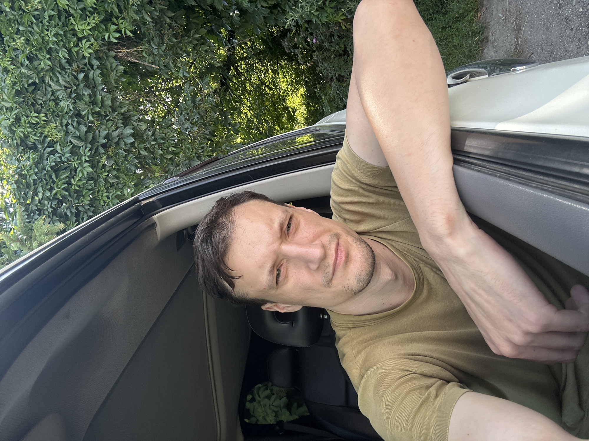 In Shatava, Ukraine, a man with short, dark hair and stubble is seated in a vehicle, with his right arm resting on the open car door. He is wearing a plain, olive-green t-shirt. The background consists of lush green foliage and trees, suggesting an outdoor, possibly rural, setting. Sunlight filters through the leaves, creating a dappled effect. The interior of the car, including the ceiling and a black headrest, is visible. The man is looking directly at the camera.