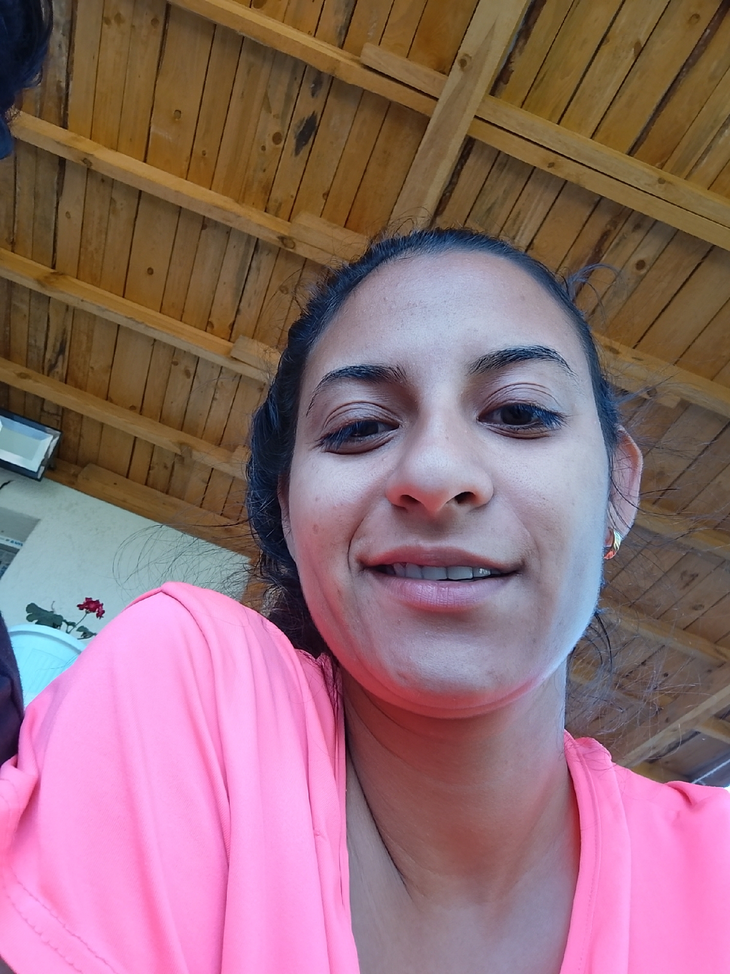 A young woman is captured in a close-up, slightly angled selfie, her face filling most of the frame. She has dark hair pulled back, exposing her ears, one of which sports a small gold hoop earring. Her skin is smooth, and she has dark, prominent eyebrows and eyelashes. Her lips are slightly parted, revealing white teeth, and she has a subtle smile. She is wearing a bright pink t-shirt, the fabric of which is visible in the lower portion of the image. The background suggests an outdoor setting, with a wooden ceiling overhead, possibly part of a covered patio or pergola. To the left, a white wall is partially visible, along with a light fixture and a small plant with red flowers. The overall lighting suggests a daytime scene. The image quality is moderate, with some visible graininess. The subject is positioned in the city of Zimnitsa, Bulgaria.