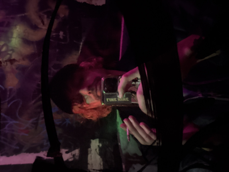 A person with reddish hair and a dark beanie is taking a photo with their phone. The phone case has "FINE NINE" on it. They are in a dimly lit area with graffiti on a nearby wall. A dark vertical pole is visible on the left, and another dark object obscures part of the view on the right. The overall scene has a slightly hazy or grainy quality.