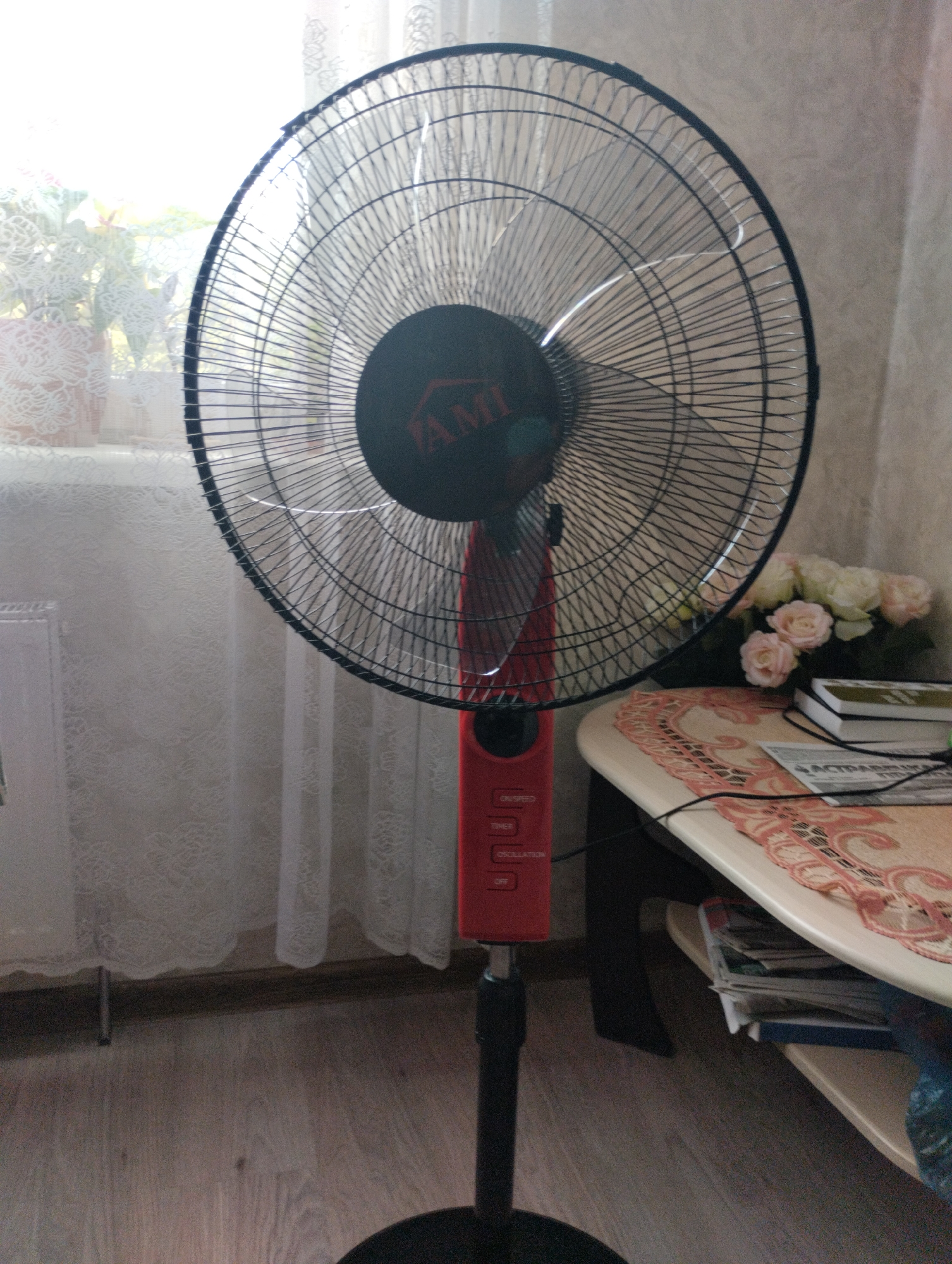 An indoor, medium shot, eye-level, full-frame photograph shows a red oscillating floor fan positioned on a wooden floor in front of a window with white lace curtains and a potted plant. The fan's black grille, with its brand logo featuring the word "AMI" in a red triangle, is the central focus. The fan's red base has several control buttons labeled "ON/SPEED", "TIMER", "OSCILLATION", and "OFF". To the right of the fan is a small, light-colored table with a decorative pink and white runner. On the table are a stack of books and a bouquet of artificial roses in shades of pink and white. Beneath the table, a stack of papers and books is visible. The environment suggests a domestic setting. No people are present in the scene. This photograph was taken in Astravyets, Belarus.