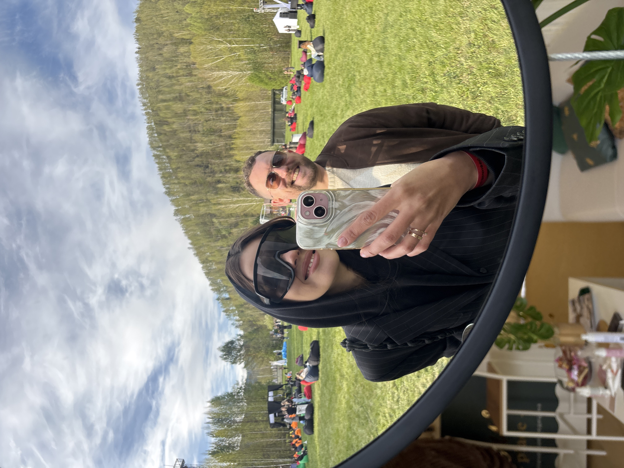 A selfie in a round mirror shows two people. The person holding the phone has dark hair, wears sunglasses, and a pinstriped jacket. A man behind them has sunglasses and a brown jacket. Reflected in the background is an outdoor setting: a grassy field with trees, people sitting in chairs or on the ground, and equipment or a structure in the distance. A portion of an indoor scene is visible on the right side of the mirror, showing shelves with objects. The image was taken near the city of Krasnoyarsk, Russia.