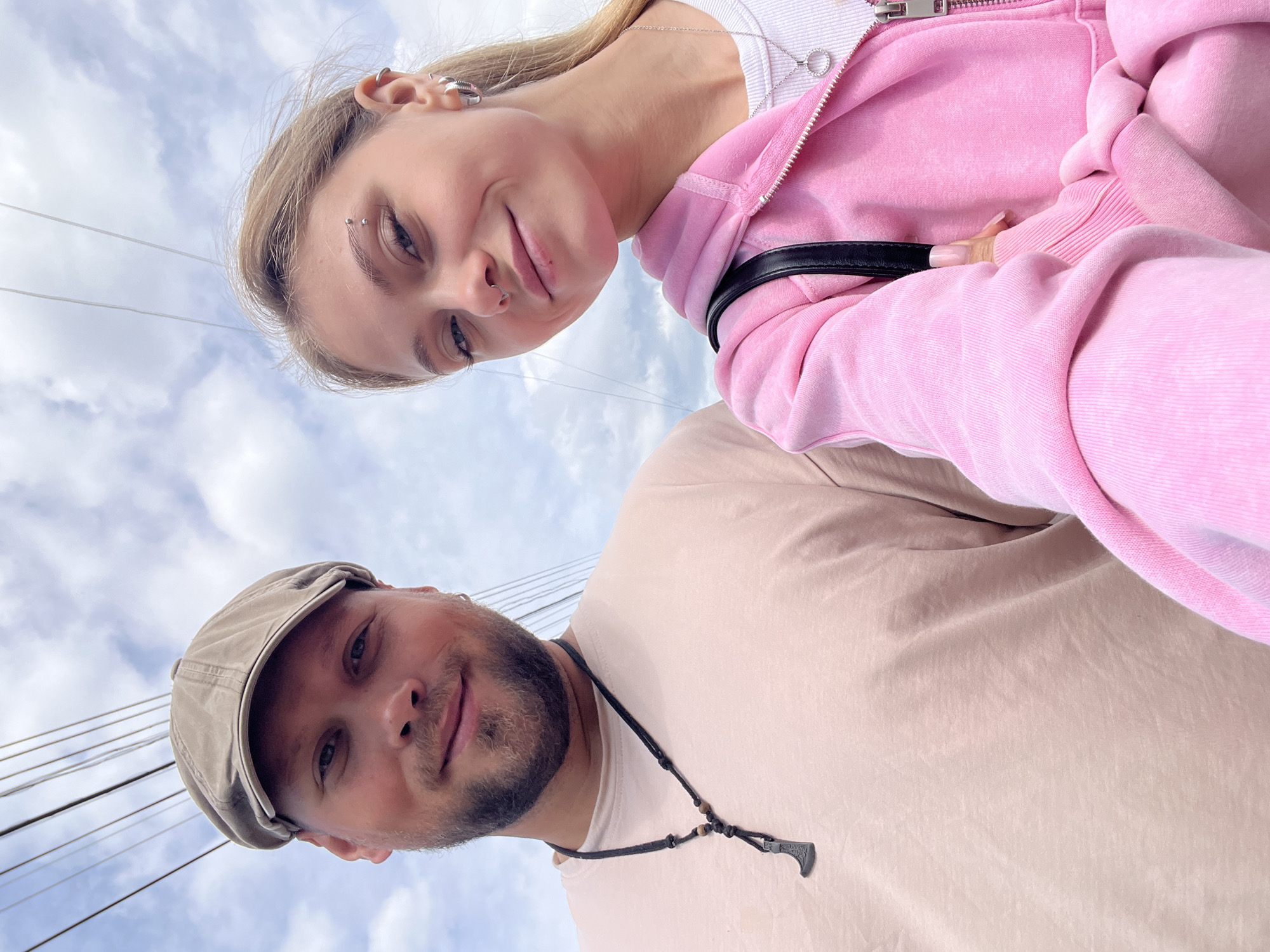 Two people pose for a selfie, angled upwards against a cloudy sky traversed by overhead wires. A woman with blonde hair, an eyebrow piercing, and a nose ring wears a pink zippered hoodie. A man with a beard and a cap wears a light tan t-shirt and a necklace with a small pendant. The location cannot be determined.