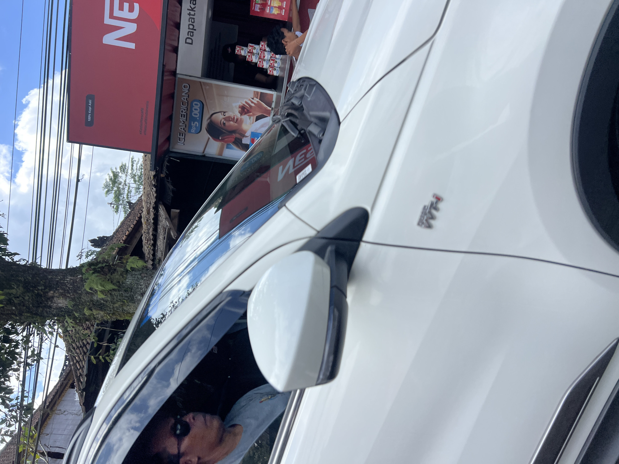 In Payangan, Indonesia, a white SUV is parked in front of a Nescafé establishment, a common sight for quick service coffee shops in the region. The driver, a man wearing sunglasses and a grey t-shirt, is visible inside the vehicle. Adjacent to the SUV, a poster advertises an "ICE AMERICANO" for Rp 5.000, featuring a young woman with headphones enjoying a beverage. The Nescafé signage is prominent, displaying the brand's logo and colors. Behind the Nescafé, traditional Indonesian architecture is visible, with tiled roofs and weathered wooden structures, typical of the local building style. A tree with lush foliage and mossy bark stands tall, its branches reaching towards the sky, creating a natural contrast with the man-made structures. Power lines are strung overhead, indicating the presence of electricity infrastructure in the area. The sky above is a mix of blue and white, with scattered clouds, suggesting a bright, possibly humid, tropical day. The scene captures a moment of everyday life in Payangan, where modern conveniences like drive-through coffee shops blend seamlessly with the traditional Balinese environment.