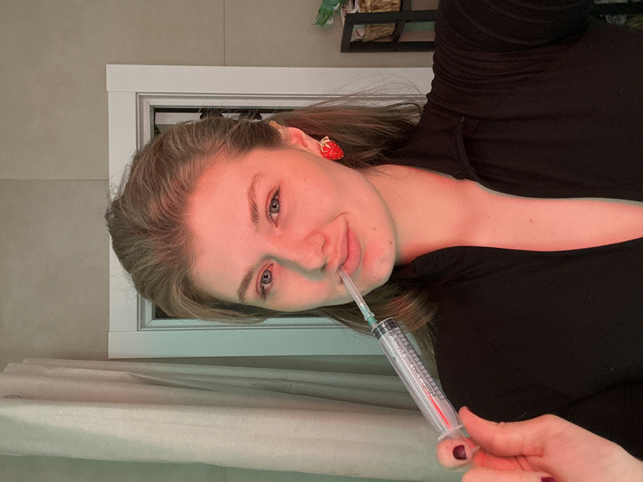 The image shows a young adult female in an indoor setting, likely a residence in Kazan, Russia. She has light skin, blue eyes, and light brown hair that is pulled back. She wears a dark, long-sleeved top with a zipper detail and a red, strawberry-shaped earring on her right ear. Her left hand, with dark-painted fingernails, holds a large, clear plastic syringe, approximately 10-20 mL in volume, featuring a green-capped needle. The covered needle tip is positioned between her lips, and she looks directly at the camera with a slight facial expression. The background consists of a light-colored tiled wall, a light-toned curtain on the left, and a white door frame on the right, beside which a dark, open shelving unit is partially visible. The scene is illuminated with colored lighting, casting red hues predominantly on the right side of her face and green hues on the left.