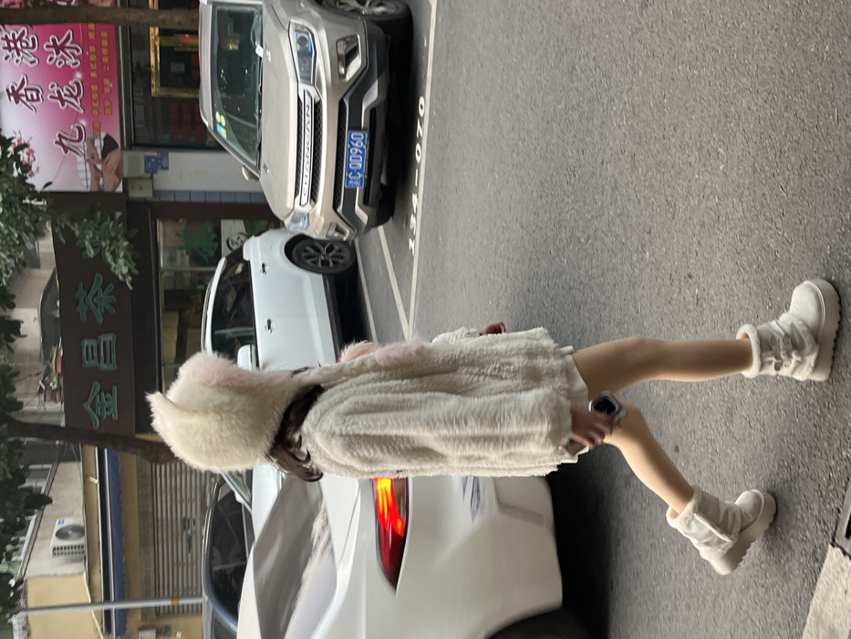 A single individual is observed walking left-to-right across an asphalt street in Jiangjin, Chongqing Municipality, China. The person, seen from the rear, is dressed in a cream-colored fluffy coat, a hooded hat featuring animal ears and pink inner accents, a light-colored garment underneath, and white platform boots with fur trim. A dark-colored smartphone is held in their right hand at hip level. Parked vehicles line the street behind the subject: a white SUV on the left and a gold Changan SUV, bearing license plate C·Q00960, on the right. Commercial building facades with signage, including "Jinchang Tea" (金昌茶) and "Kowloon Ice" (九龙冰), are visible behind the vehicles. Parking spot numbers "134-070" are marked on the pavement.
