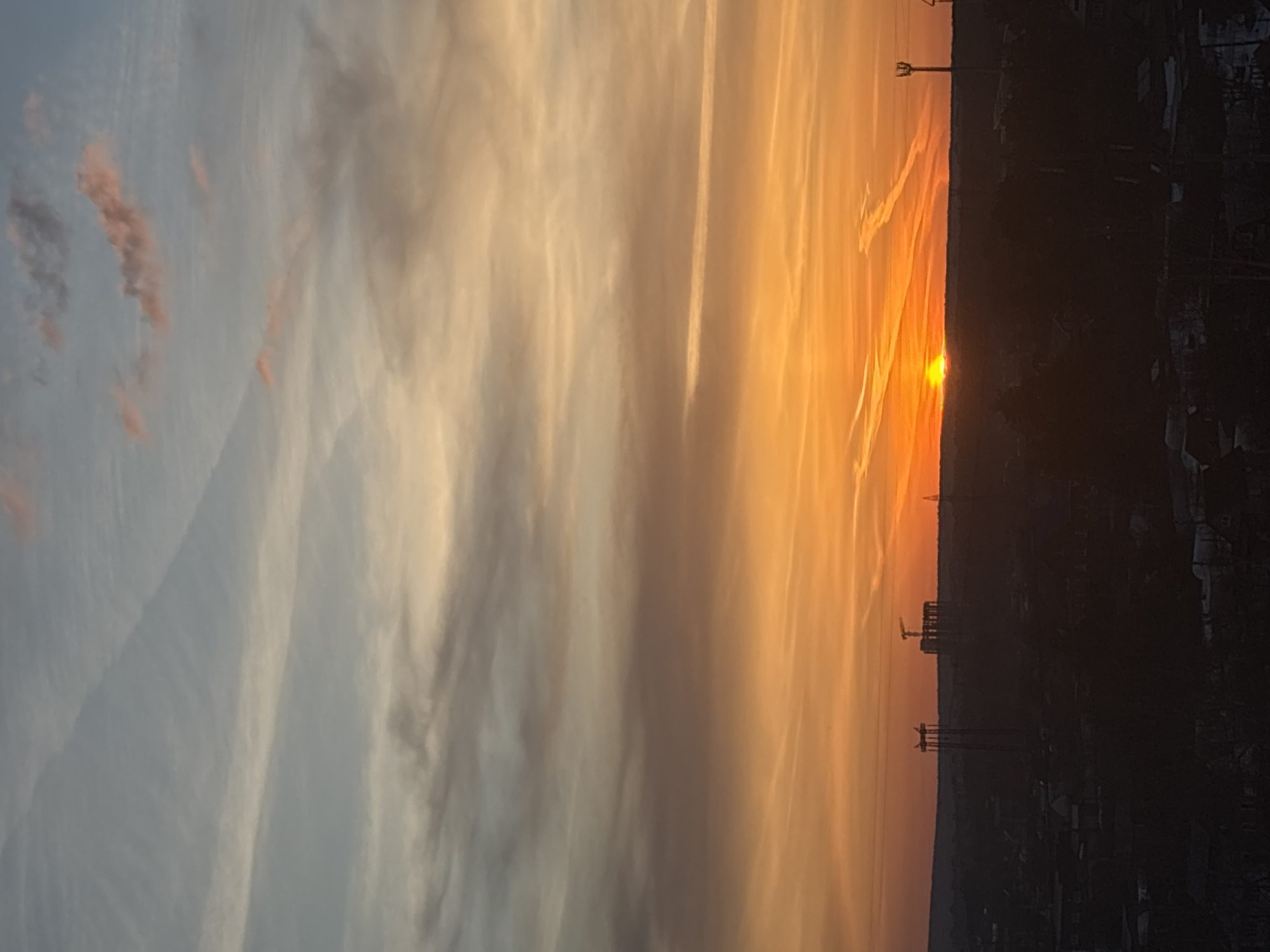 The image captures a sunrise in the city of Chaikovsky, Russia. The sky transitions from blue with hints of pink clouds on the left to a vibrant orange near the horizon on the right. The sun is partially obscured by the dark silhouette of buildings, trees, and industrial structures, including cranes and power lines. The foreground is a dark, indistinct cityscape. The image is rotated 90 degrees clockwise. Contrails are visible in the sky. No people are directly visible.