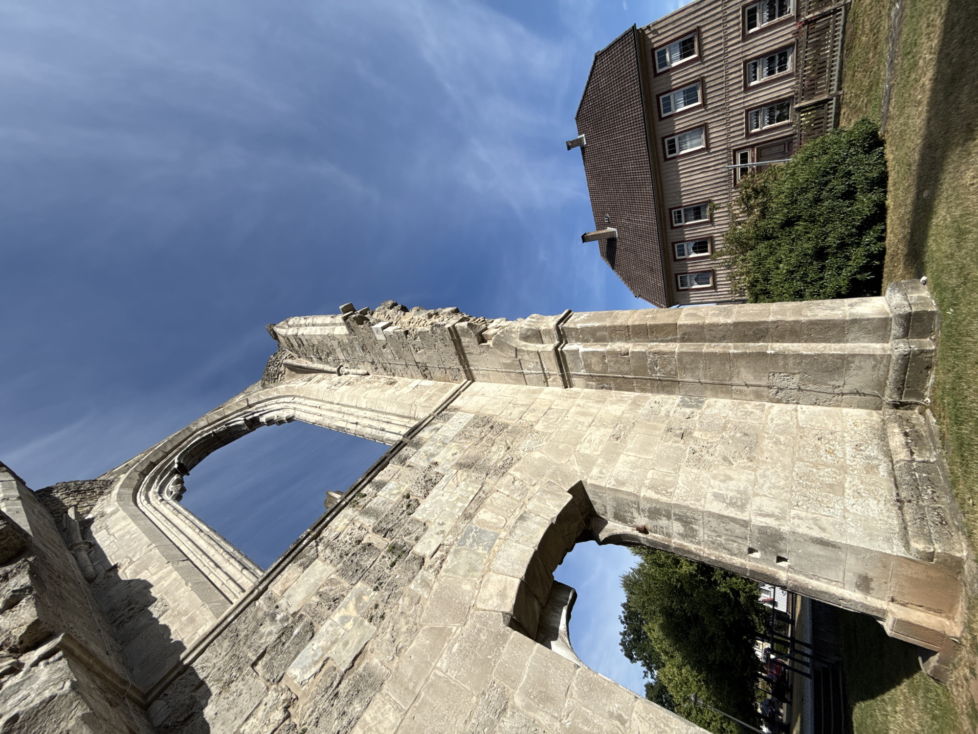 The image, taken in Walkenried, Germany, captures a section of the historic Walkenried Abbey ruins, a prominent historical site in the town. In the foreground, the weathered stone structure of the abbey's wall dominates the left side of the frame, showing architectural details like arched windows and rough-hewn blocks. The sky above is a vibrant blue with scattered white clouds. To the right of the ruins, a traditional wooden-clad house with a tiled roof is visible, its windows reflecting the sunlight. A dense green bush grows in front of the house, and a wooden fence separates it from the grassy area. The overall impression is one of historical preservation and a peaceful, sunny day in the town.