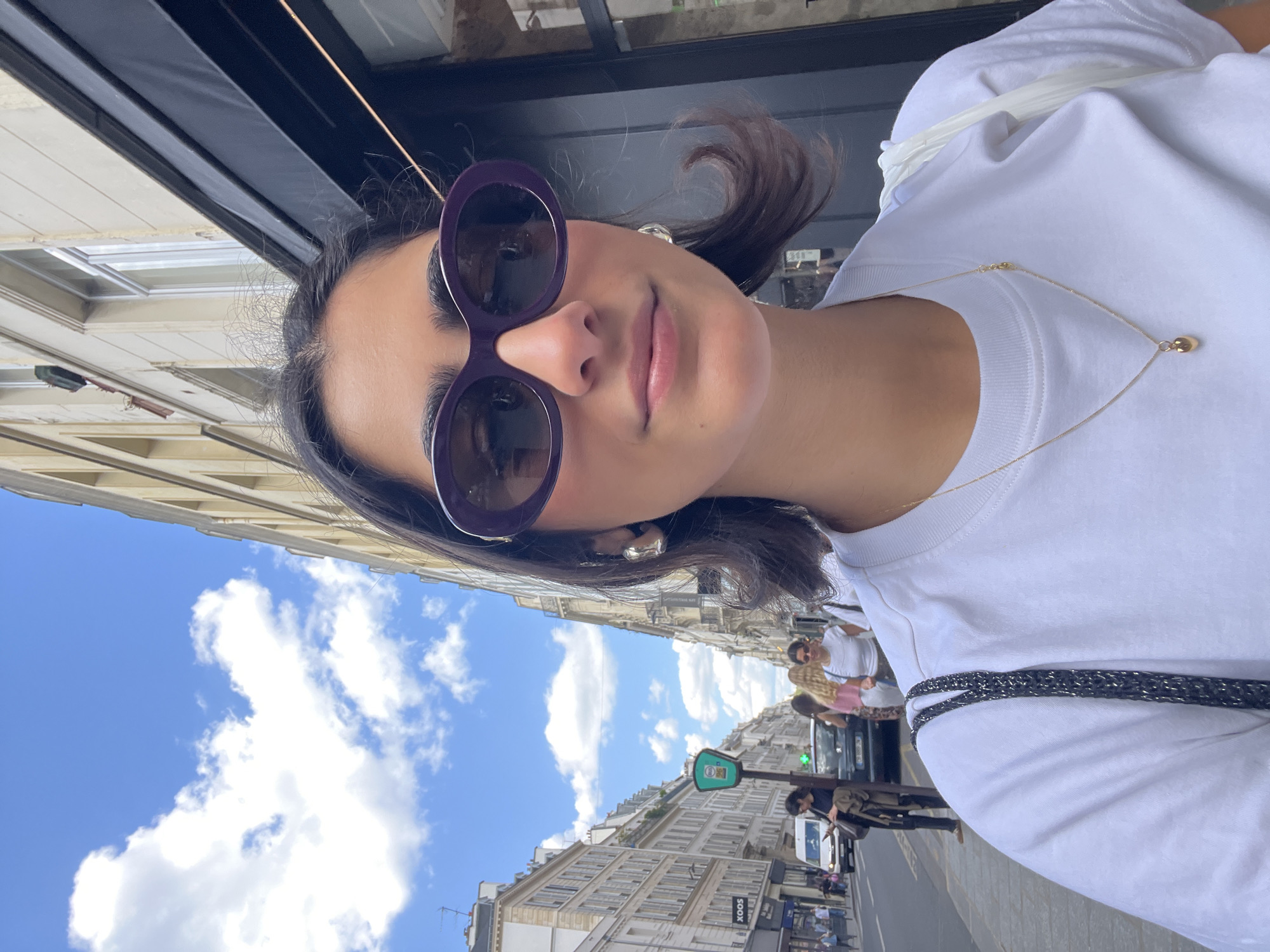 A woman is taking a selfie in Paris, France. She wears large purple sunglasses, a white t-shirt, and a gold necklace. A black bag strap is visible on her shoulder. The streetscape shows typical Parisian architecture with buildings lining the street. People are walking along the sidewalk and cars are on the road. The sky is blue with scattered white clouds. A green street sign is visible near a crosswalk.
