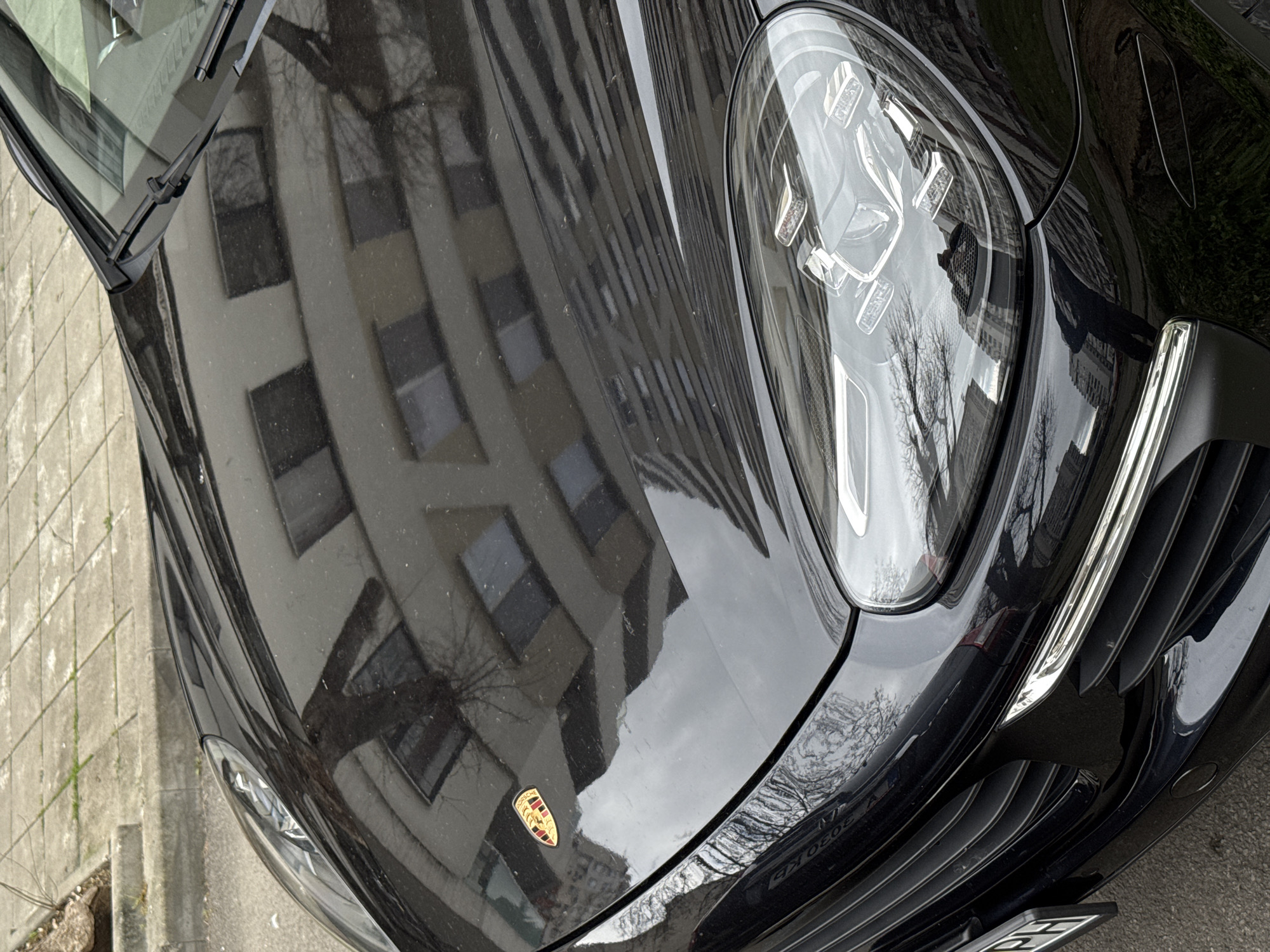 A black Porsche is parked on a street in Burgas, Bulgaria. The hood reflects a nearby building. The car's headlights, Porsche emblem, and part of the license plate are visible. The surrounding environment includes a sidewalk and possibly a tree as reflected in the car's paint.