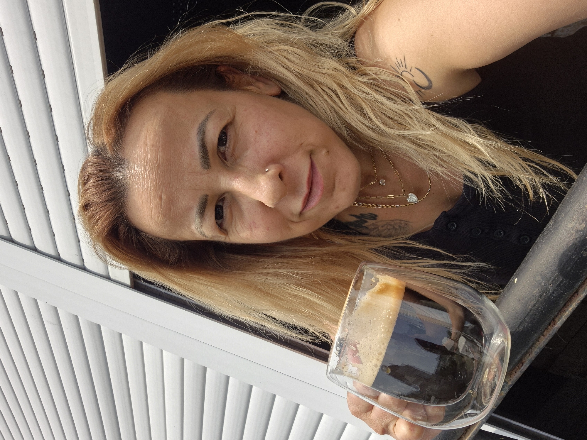 A woman with blonde hair and tattoos holds a glass of coffee in front of a window with white blinds. She is wearing a black top and multiple necklaces. Her face is turned towards the camera, and she is smiling. The scene appears to be outdoors.
