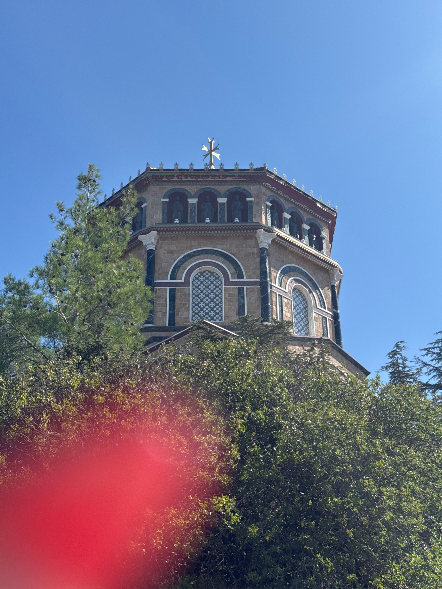 In the city of Mylikouri, Cyprus, a low-angle, medium shot captures the upper portion of a multi-story, octagonal building, likely a church, against a clear, bright blue sky. The building's facade is constructed of light-colored stone with decorative bands of dark green and red. Several arched windows, each with intricate white patterned glass, are visible. The top of the structure is adorned with a series of small, white decorative elements and topped with a prominent golden cross. The building is partially obscured by lush green foliage from trees and bushes in the foreground. A large, out-of-focus red circular shape is present in the lower left quadrant of the image, suggesting a lens flare or a physical obstruction.