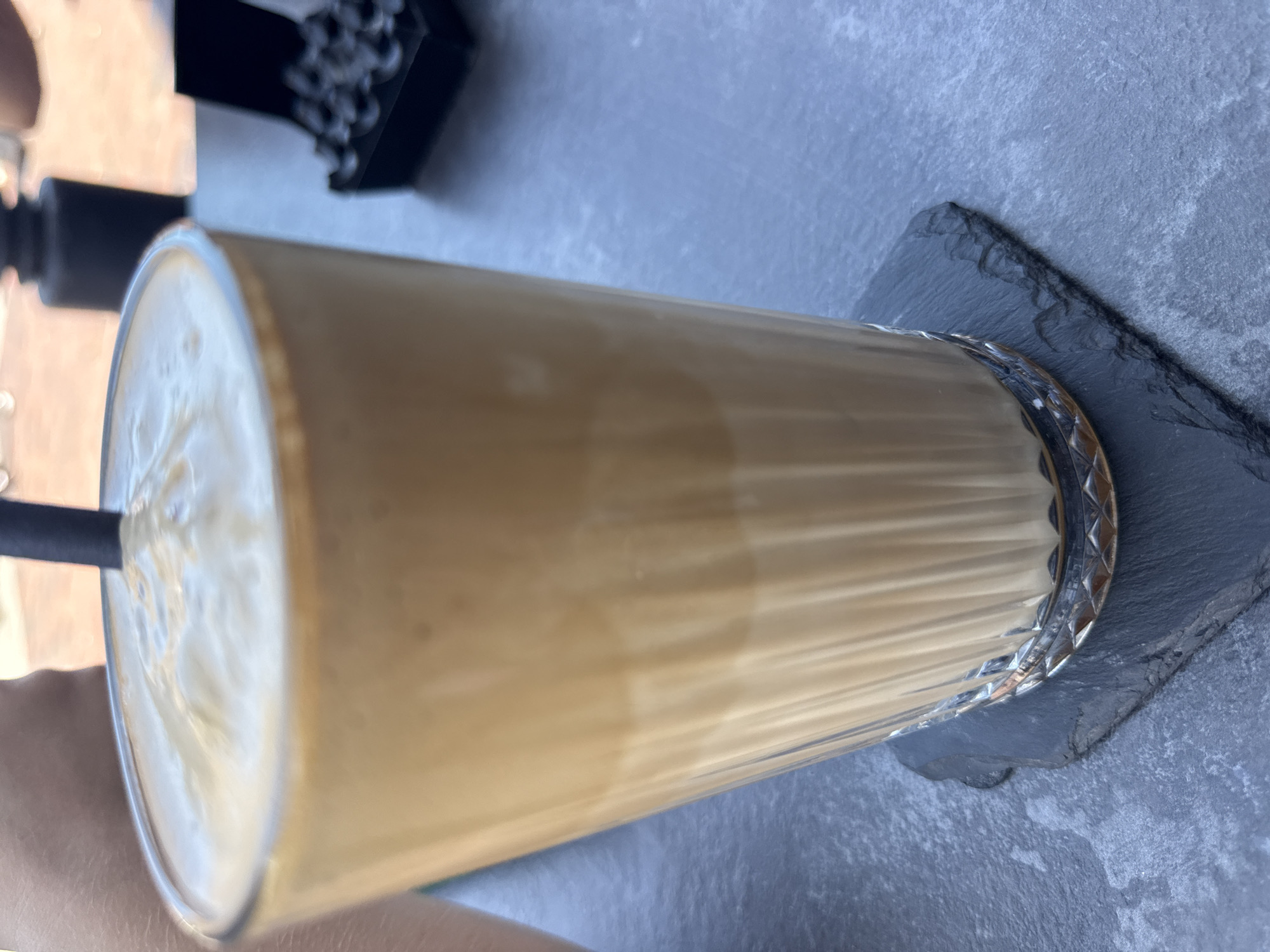 A tall glass of iced coffee with a frothy top sits on a dark slate coaster on a gray, textured table. A black straw is visible in the drink. In the background, out of focus, are a black object and a black box filled with small, dark beads. The image is taken from a slightly elevated angle.