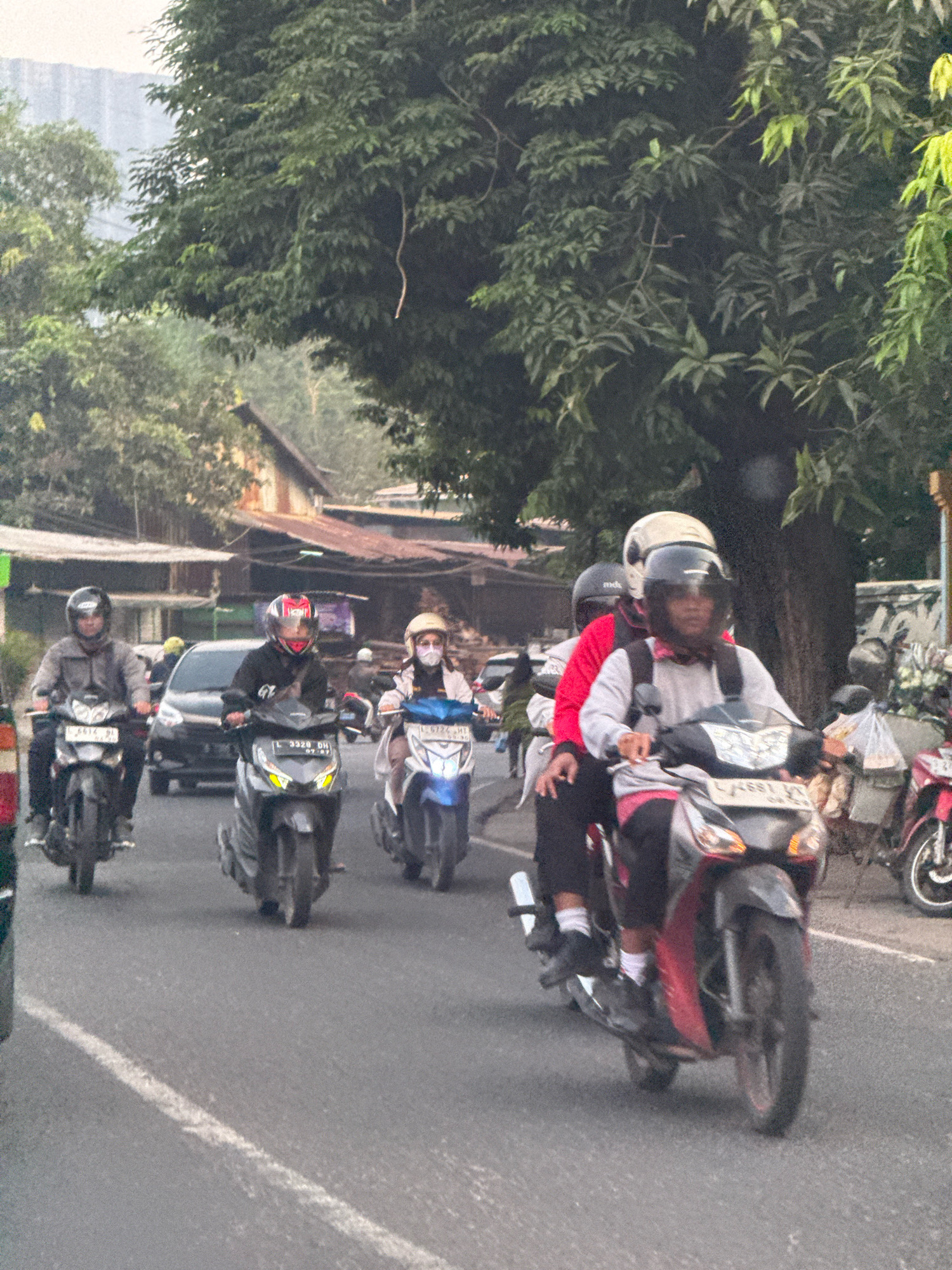 A medium-shot, eye-level, vertical, outdoor, daylight, medium-resolution, color photograph depicts a street scene in Surabaya, Indonesia, with several motorcycles traveling on the asphalt road. The motorcycles are moving from left to right in the frame, and some are ridden by multiple people. The road is bordered by trees and buildings, suggesting an urban or semi-urban environment. The lighting is overcast, creating soft shadows.
In the foreground, a motorcycle with two riders is visible. The driver wears a white helmet and a grey long-sleeved shirt with pink accents, and is looking forward. The passenger behind them wears a black helmet and is dressed in dark clothing. The motorcycle is red and black. To their left, another motorcycle is being ridden by a person wearing a grey jacket and a black helmet. The motorcycle is black.
Further back, a motorcycle with a red and black helmet is being driven by a person in a black jacket with white lettering. This motorcycle is dark grey. To the right of this, another motorcycle has a rider wearing a beige helmet and a white jacket, and a face mask. This motorcycle is white and blue. Several other motorcycles are partially visible in the background.
A dark-colored car is visible behind the motorcycles in the middle ground. The background features buildings with corrugated metal roofs and a dense canopy of green trees, some of which appear to be large and mature. The sky is a hazy grey. The road is marked with a white line. The overall impression is of a busy street with moderate traffic.