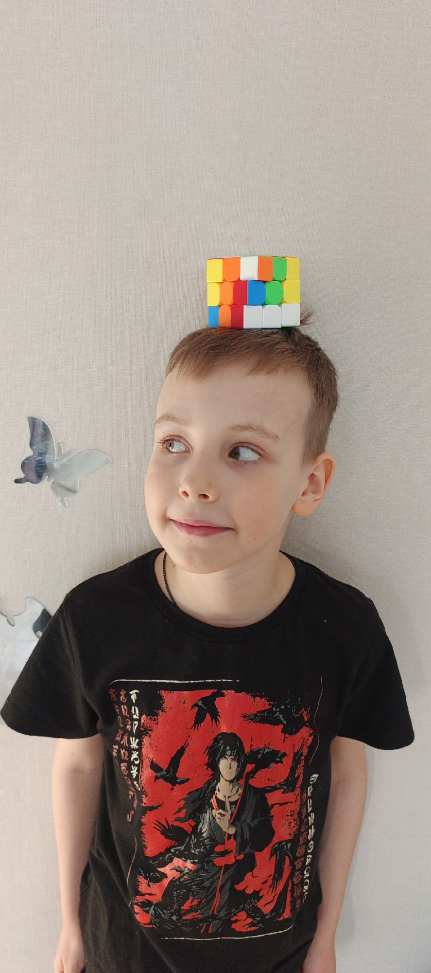 A young boy with short brown hair, fair skin, and brown eyes balances a Rubik's Cube on his head. He is wearing a black T-shirt with a red graphic featuring a character surrounded by ravens. The background is a beige wall with a silver butterfly decoration to the left of the boy's head. The photo appears to have been taken in an indoor setting. I cannot determine the location of Chaikovsky, Russia from this image.