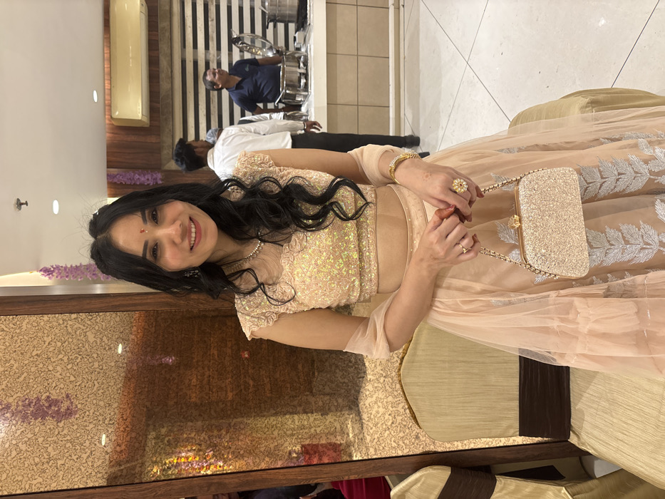 A woman smiles, adorned in a peach-colored lengha with a sequined bodice. She holds a sparkly gold purse by its chain handle. Her hair is black and wavy, and she wears jewelry: a necklace, earrings, a bracelet, and two rings. Two men in casual clothing are visible in the background, near a buffet setup with warming trays. The photo is taken at a slight angle, seemingly in a restaurant or event venue.