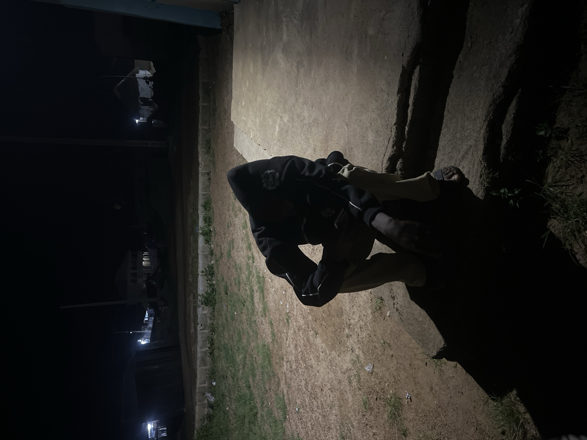 The image shows a person lying on the ground at night, likely in Durumi, Nigeria. The person is wearing a dark hooded jacket and appears to be asleep or resting. The scene is dimly lit, with shadows cast by an unseen light source. In the background, some buildings and streetlights are visible, suggesting an urban or semi-urban setting. The ground is a mix of dirt and grass, with some litter scattered around. The overall mood is quiet and peaceful, with no apparent activity other than the person's repose. The time of day is clearly nighttime due to the darkness and artificial lighting. No visible text is present in the image.