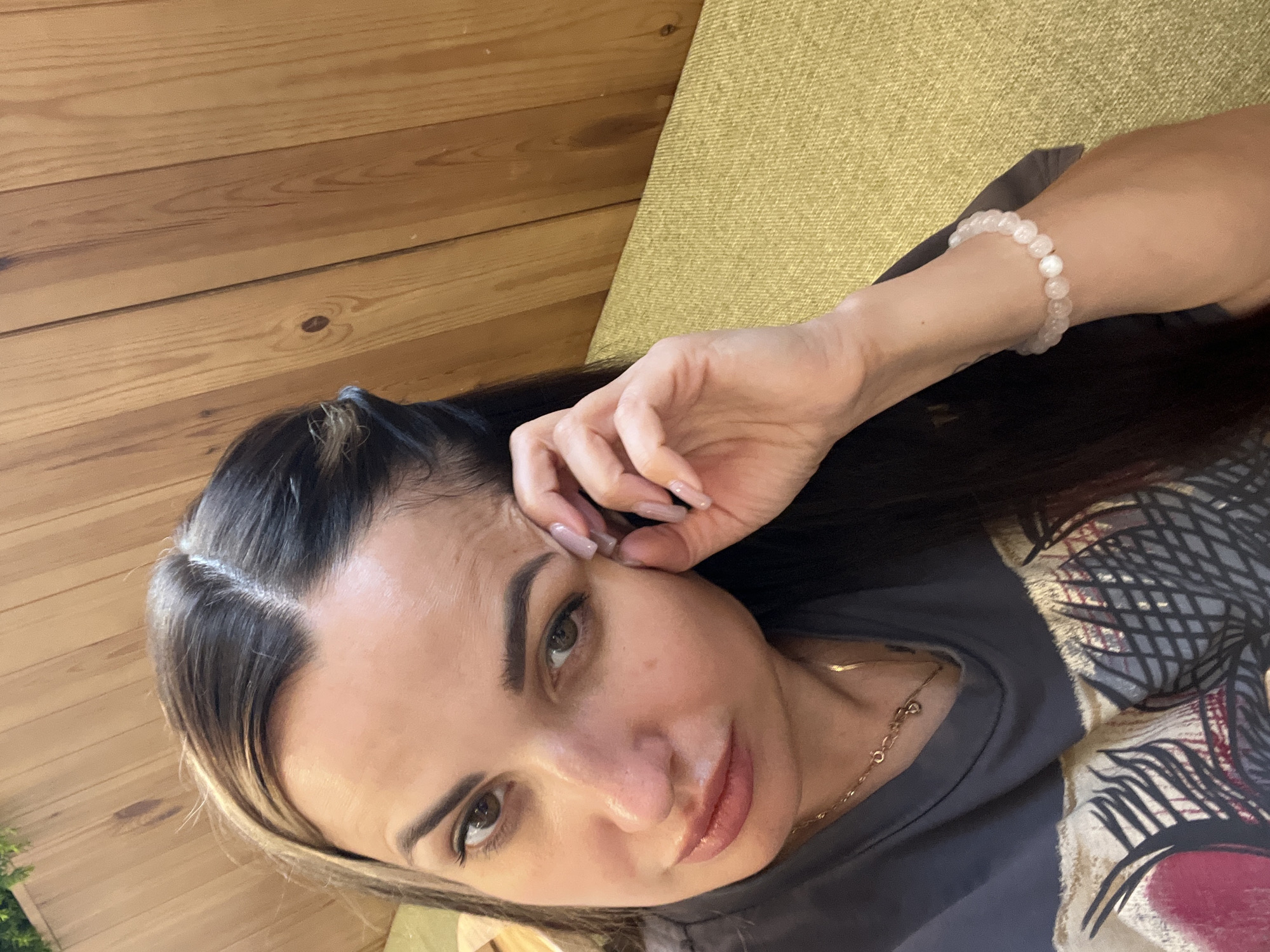 A woman is captured in a close-up, self-portrait style photograph. She is seated, leaning towards the camera, with her right hand gently touching her temple. Her gaze is directed towards the lens, and her lips are slightly parted. She has dark, long hair that is parted in the center, with a few strands swept back by a hair clip. She is wearing a dark gray t-shirt with a graphic print on it, and a delicate gold necklace. On her left wrist, she wears a bracelet made of rose-colored beads. The background consists of a textured, light brown wooden wall and a yellow upholstered seat. The lighting is soft, casting subtle shadows on her face. The overall impression is that of a casual, personal moment.