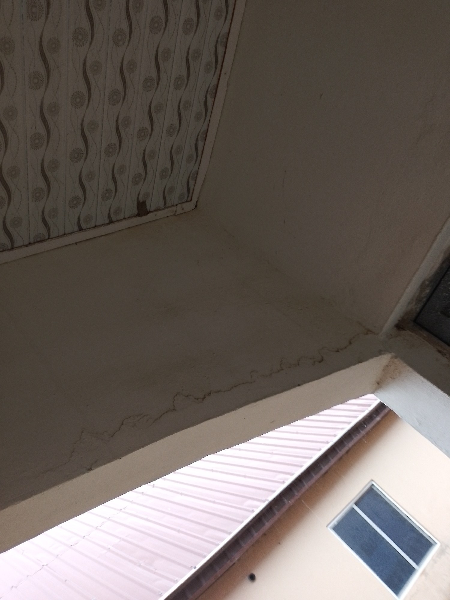 The image depicts an exterior view of a building, likely in Jauro Yinu, Nigeria, from a low-angle perspective looking upwards. The foreground features a section of a textured, beige-colored overhang. Below this overhang, a pink corrugated metal roof is visible, with a brown gutter running along its edge. Further in the background, a light beige building with a dark-framed window is partially seen. The upper portion of the image reveals a ceiling with a patterned design, consisting of wavy lines and circular floral motifs in shades of grey and off-white. There are no people or discernible actions taking place in the scene. The environment appears to be an outdoor space adjacent to a building.