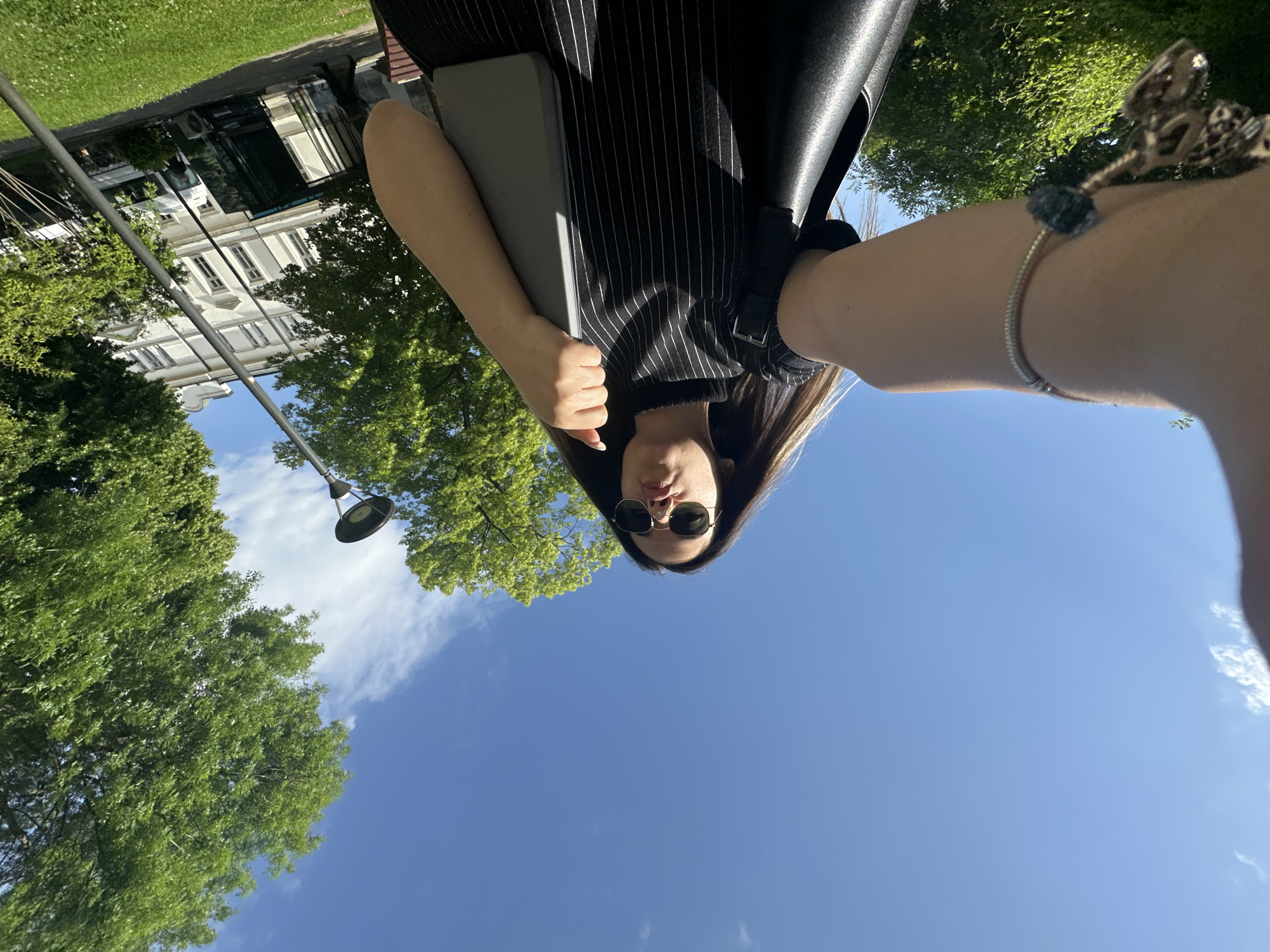 A woman in Sofia, Bulgaria, takes a selfie outdoors on a sunny day. She wears sunglasses and a black pinstripe dress or shirt, holding a grey laptop or book. She also wears a silver bracelet. Trees and a blue sky fill the background. A white building with multiple windows is visible in the distance, and a lamppost appears on the left. The image is rotated counter-clockwise, so the sky is at the bottom and the woman's head is near the center.