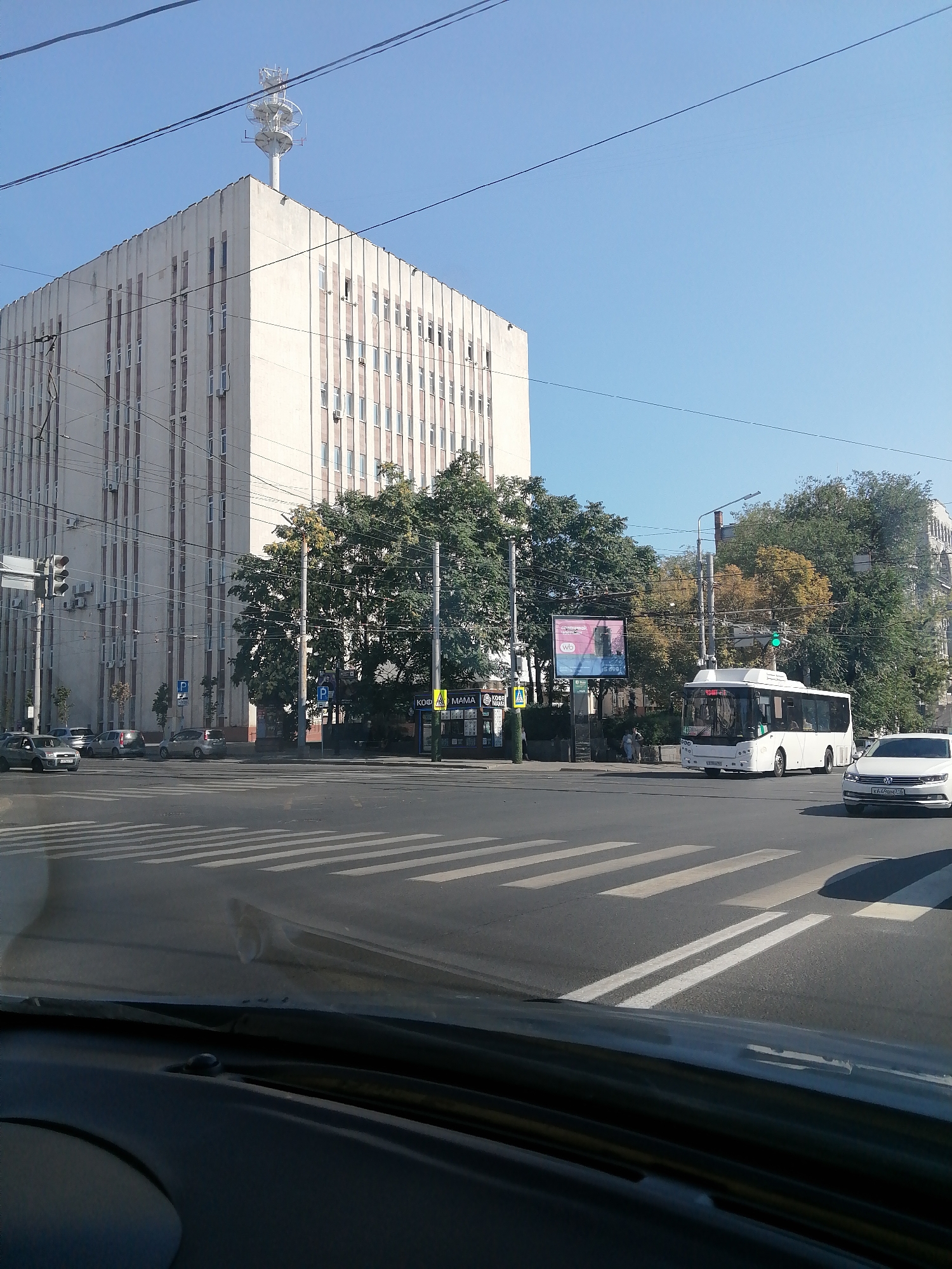 The image depicts a street scene in Rostov-on-Don, Russia, with a large, multi-story administrative building dominating the left side of the frame. This building, characterized by its light-colored facade and vertical window arrangement, features a prominent telecommunications tower on its roof. A traffic intersection with a pedestrian crossing is visible in the foreground. A white bus, identified by its license plate as likely Russian, is in motion on the right side of the intersection, while a white Volkswagen sedan is stopped at the crosswalk. Several parked cars are also present on the street. The environment is urban, with trees lining the sidewalk and utility wires crisscrossing the sky. The sky is clear and blue, indicating daytime.