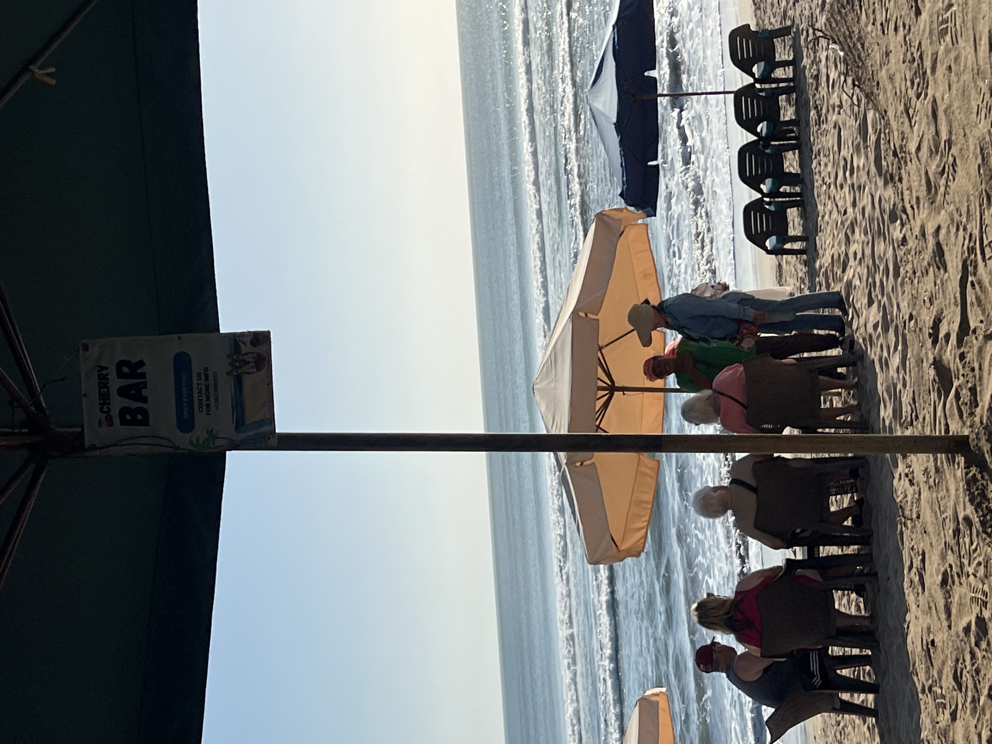 A group of people are sitting at tables under umbrellas on a sandy beach in Kuta, Indonesia, facing the ocean. The ocean has moderate waves, and the sky is clear and bright. A sign for "CHERRY BAR" hangs from a pole. Several lounge chairs are set up on the beach.