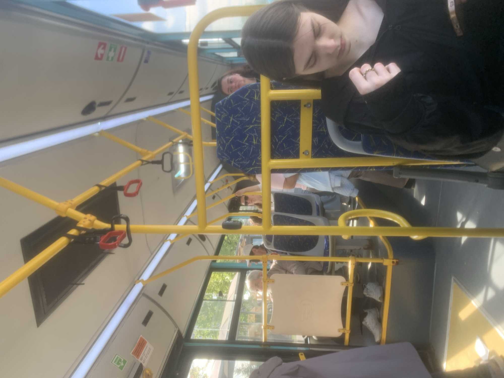 The image captures the interior of a public bus or trolleybus, possibly in motion during the daytime on a sunny day. The perspective is angled, looking towards the front and side of the vehicle.
In the foreground, a young woman with dark hair, wearing a black top and a ring on her finger, appears to be resting or sleeping, leaning her head against a bus seat. Her eyes are closed. Behind her, another passenger is partially visible, also seated.
The bus interior is characterized by bright yellow handrails and vertical poles that crisscross the frame, providing support for standing passengers. Some of these poles have red loop handles attached. The seats are upholstered in a blue fabric with a light-colored, abstract pattern. Several other passengers are visible further down the bus aisle; one elderly person with white hair and a cap is seen through a side window.
Sunlight streams through the large windows, illuminating the interior and revealing greenery and blurred structures outside, indicating movement. The floor is grey, and some white shoes are visible near the back of the bus. Overhead panels are light grey, featuring linear lighting fixtures and various signs.
Visible text and symbols include:
- On an overhead panel, there's a green symbol of an arrow pointing left and a red symbol of two figures (possibly indicating a gender-neutral restroom or changing room, or an emergency exit). Another red symbol, possibly an emergency brake sign, is also present.
- A smaller green emergency exit sign with a running person and an arrow is visible on a panel further back.
- Other small, unreadable text blocks are present on various panels.
The overall atmosphere suggests a typical commute, with passengers either observing the outside or resting during their journey. The bus structure and design are consistent with public transport vehicles found in many European countries, including Russia.