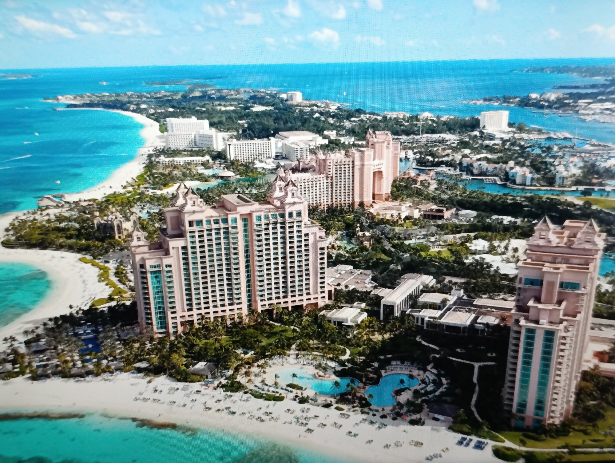 This is an aerial, wide-angle shot of a tropical resort complex situated on a coastline. The dominant features are several large, peach-colored hotel buildings with distinctive architecture, including arched windows and tiered roofs. One prominent building is a very tall, rectangular tower with numerous windows, some of which are accented with turquoise glass. Another large structure features a grand, arched entryway.
The resort grounds are lush with greenery, including numerous palm trees, and are dotted with swimming pools, some of which are shaped like aquatic animals. There are outdoor lounge chairs and umbrellas scattered on white sandy beaches that meet the bright turquoise ocean. The water appears clear and calm, with small boats visible in the distance. The sky above is blue with scattered white clouds.
The scene is not located in Dubăsari town council, Moldova. Based on the architecture and tropical environment, this appears to be a resort in a warm, coastal region, possibly the Caribbean or a similar destination.