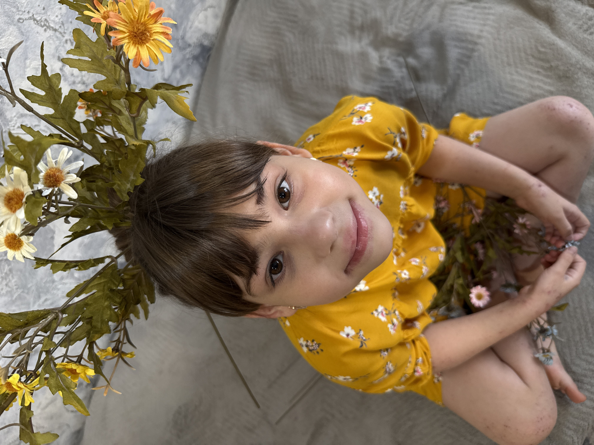 A young girl with brown hair and eyes is sitting cross-legged on a gray surface, possibly a blanket or sheet. She is wearing a yellow dress with a small white floral pattern. She looks up at the camera and smiles slightly. Her head is adorned with a floral crown of yellow and white flowers, with some flowers also being held in her hands. The background appears to be a mottled gray or white.