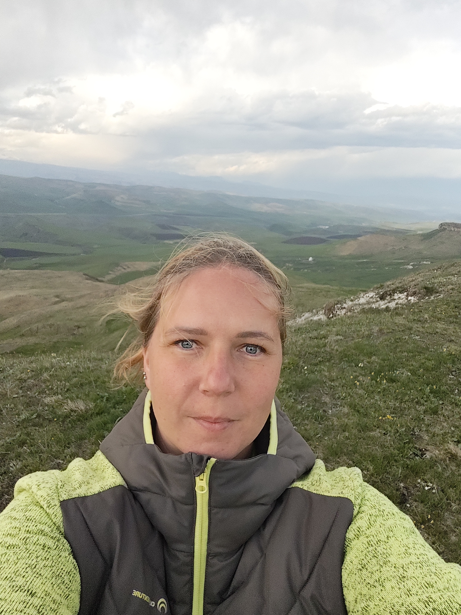 A woman is taking a selfie outdoors, likely from a high vantage point. She has fair skin, blonde hair, and blue eyes. She is wearing a green and gray zip-up jacket.
The background consists of rolling green hills and fields. In the distance, there are some darker areas that could be forests or shadows from clouds. The sky is overcast with white and gray clouds.
Given the information, the location is Kichmalka, Russia. The elements in the image seem consistent with the environment.