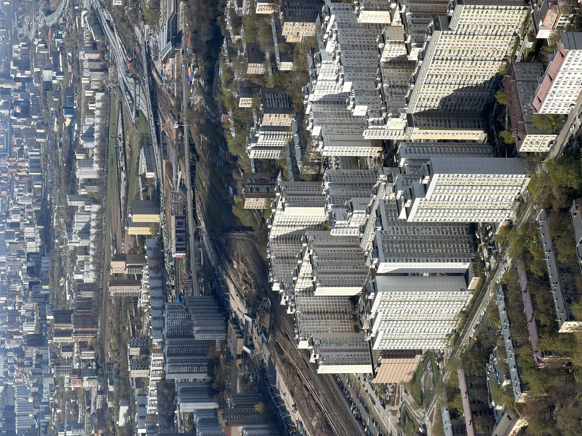 This is an aerial photograph of a residential area in Khimki, Russia, characterized by dense clusters of multi-story apartment buildings. The buildings exhibit a repetitive, modular design. A railway track runs through the area, creating a dividing line. Green spaces appear intermittently between the buildings, suggesting parks or tree-lined streets. The overall impression is one of urban density and orderly development.