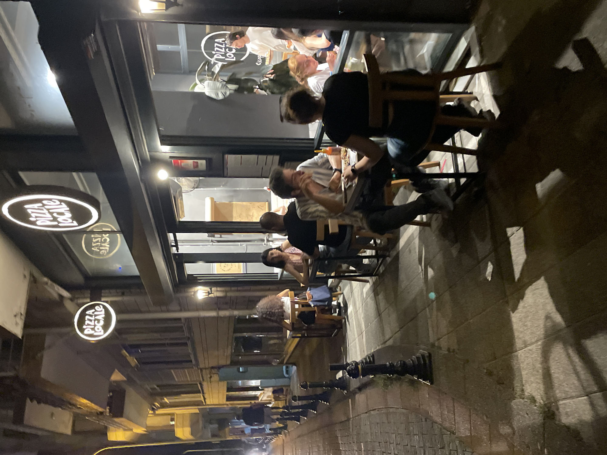 The image depicts an outdoor seating area of a restaurant called "Pizza Locale" on a street at night in Kadıköy, Turkey. Several individuals are seated at tables, engaged in dining and conversation. The restaurant has large windows, and the interior is visible, with more patrons and staff. A sign for "Pizza Locale" is illuminated and mounted on a pole outside the establishment. The street is paved with cobblestones and has a sidewalk with decorative bollards. The scene suggests a lively evening atmosphere at a popular eatery.