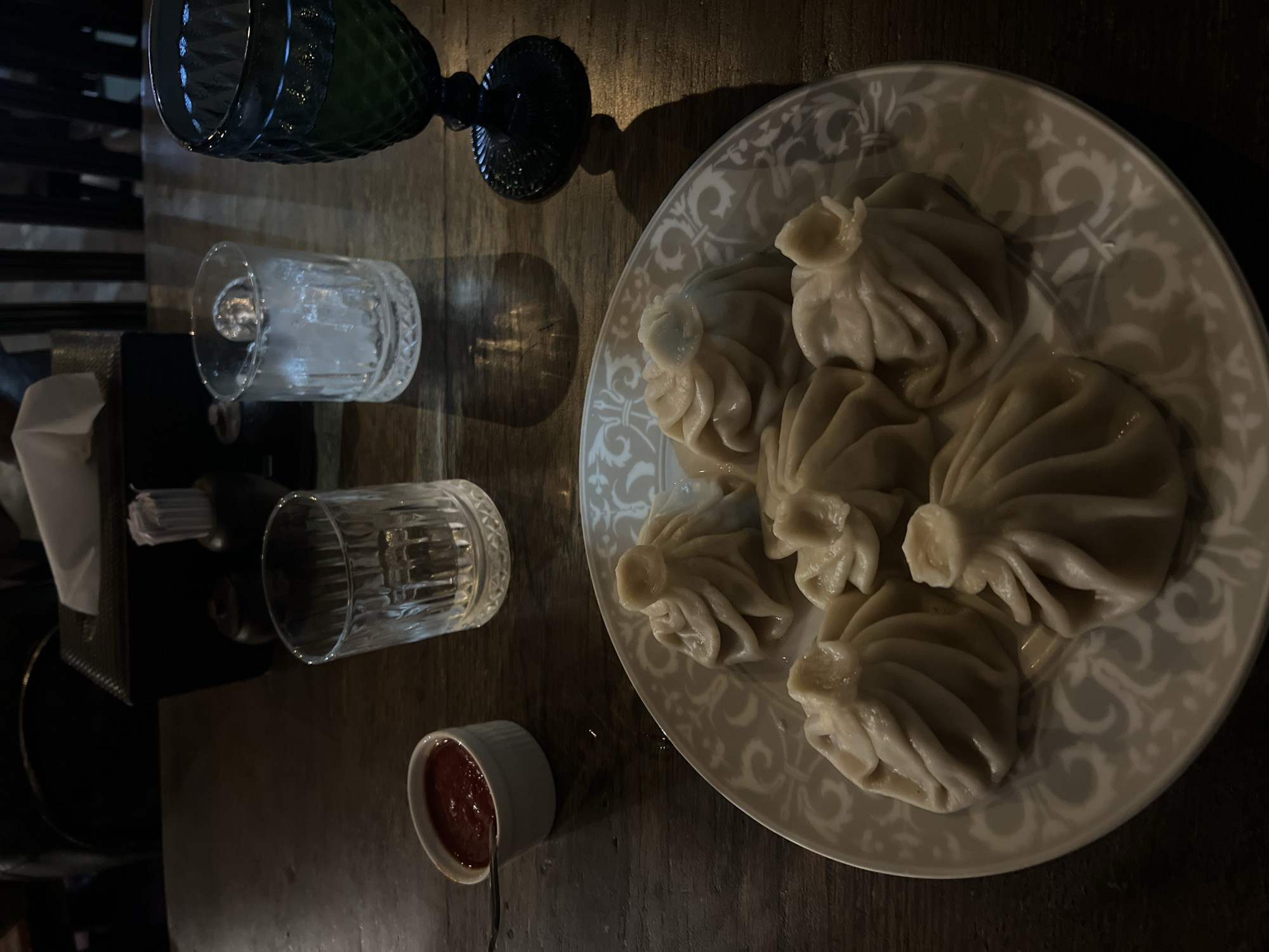 The image depicts a plate of khinkali (Georgian dumplings) arranged on a white decorative plate. Beside the plate are two textured glasses, one partially filled with a clear liquid, and a small white bowl filled with a red sauce. A decorative green glass is positioned near the top of the image. The scene is set on a dark wooden table. The lighting suggests an indoor restaurant setting. There is no discernible indication that the image was taken in North Kuta, Indonesia. There are no people visible in the image.