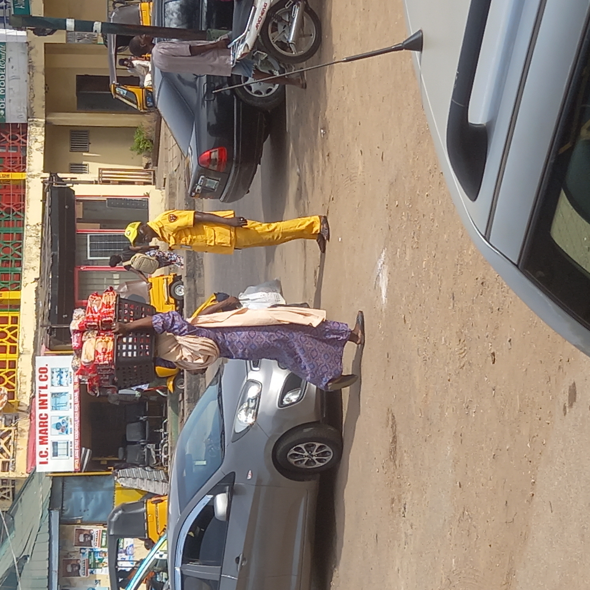 This vertically oriented image captures a lively daytime street scene in Sardauna, Nigeria. The ground is a light brown, dusty road with visible tire tracks, suggesting active vehicle movement.
In the foreground, a silver hatchback car is visible on the right side. On the road, a person dressed in a bright yellow uniform, including a cap, shirt, and trousers, walks forward, possibly a traffic controller or security official. Further to the left, a woman in a purple patterned dress and a light-colored head wrap skillfully balances a large, dark-colored crate filled with various packaged goods, predominantly red and white, on her head as she walks.
In the middle ground, a dark grey or black sedan is parked or moving slowly. Near it, a white motorcycle with "JIANJING" visible on its side is parked. A thin black pole extends diagonally across the upper part of the image, possibly a street light or utility pole.
The background reveals a row of commercial buildings or shops on the left side of the street. One prominent sign reads "I.C. MARC INT'L CO." vertically, featuring graphics of various products. A partially visible sign above it appears to say "DE MODE OUTFIT". Yellow tricycles, commonly known as "keke Napeps," are present, with one visible near the shops and another smaller one further back. Several other people can be seen in the background, engaged in various activities, indicating a busy urban environment. The bright natural lighting suggests a clear day.