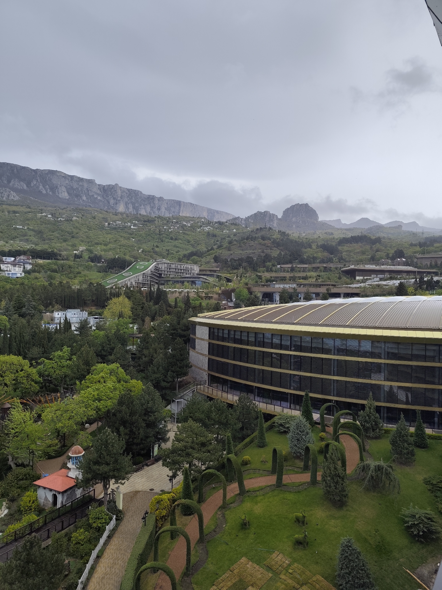 The image shows an overcast view of Симеизский поселковый совет, Ukraine. Prominently featured is a large, modern building with a curved roof and gold trim. Below the building is a meticulously manicured garden with arched pathways and topiary. In the background, a mountain range is visible, partially obscured by low-hanging clouds. Other buildings are scattered throughout the greenery in the midground. The sky is uniformly gray, indicating a cloudy or overcast day.