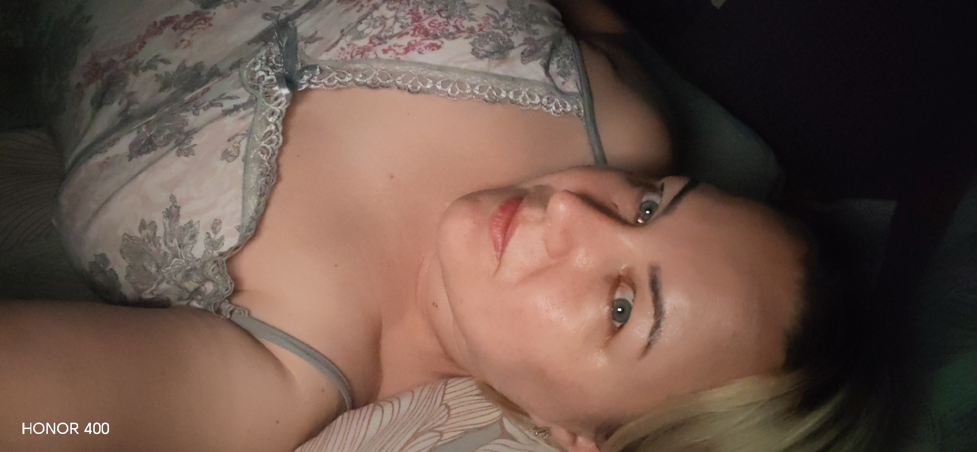 An adult woman is captured in a close-up portrait, lying down indoors in what appears to be a bedroom or living room in Noviy, Russia. She has light blonde or light brown hair and striking light-colored, possibly green or blue-green, eyes that gaze directly at the viewer. Her lips are tinted with a red or pink color, and her expression is calm and neutral, with a slight hint of a smile.
She is wearing a light-colored, possibly white or cream, nightgown or camisole with a delicate floral pattern featuring grey, pink, and reddish tones. The garment has thin straps and is trimmed with intricate white or light grey lace along the neckline. The woman's bare chest and shoulders are visible, showing clear skin.
The lighting in the image suggests an indoor setting, possibly during the evening or night, with some areas of the background being quite dark, contrasting with the illuminated subject. Part of a bed or sofa with striped or patterned bedding/cushions is visible beneath and around her. In the bottom left corner of the image, the text "HONOR 400" is visible, indicating the device used to capture the photograph.