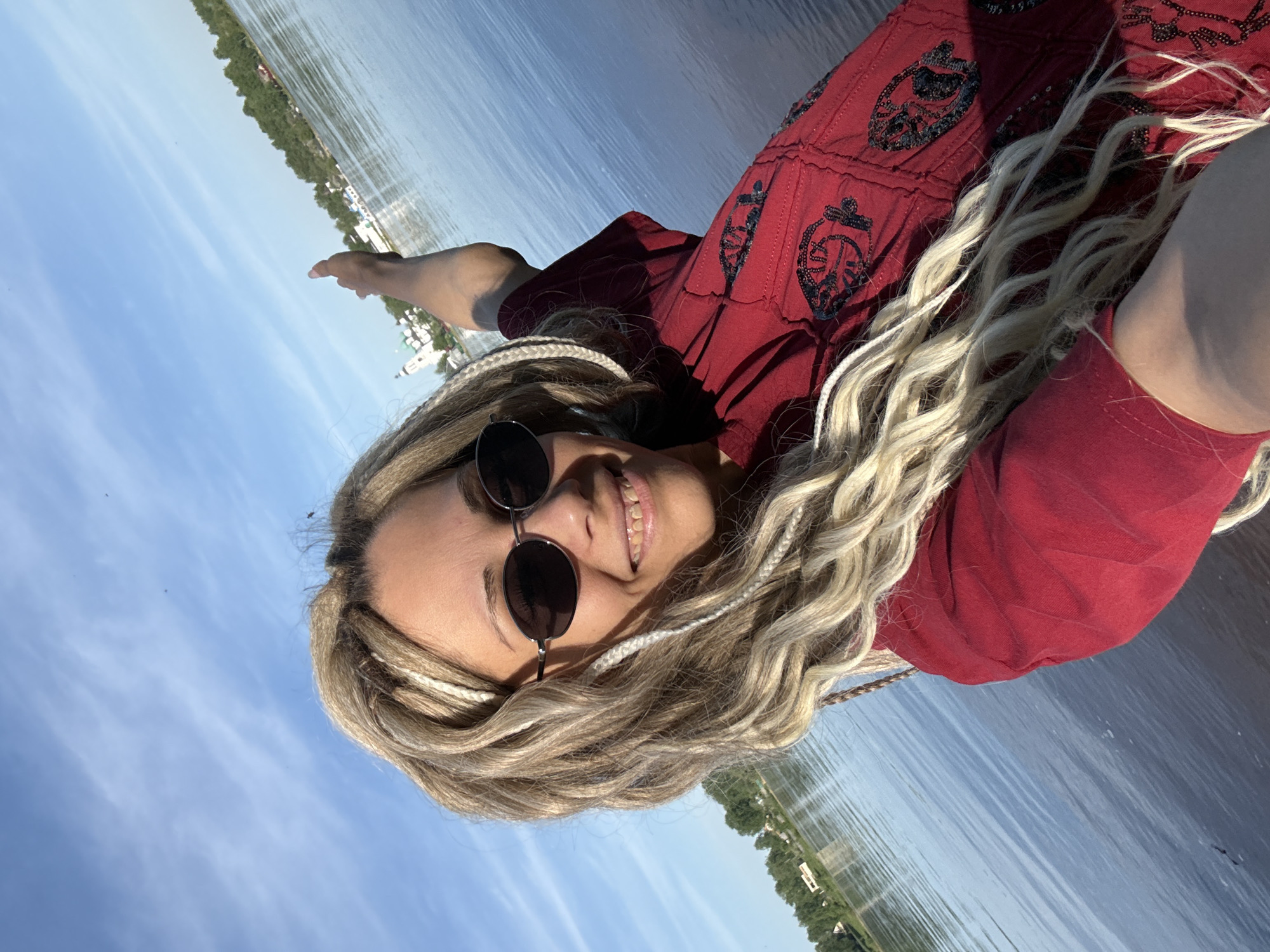 A woman with blonde dreadlocks and round sunglasses smiles at the camera. She wears a red dress with embroidered details. The background shows a body of water, likely a river or lake, with the sky visible as well. A small stretch of land with buildings is visible in the distance. The location is Unknown, Unknown.
