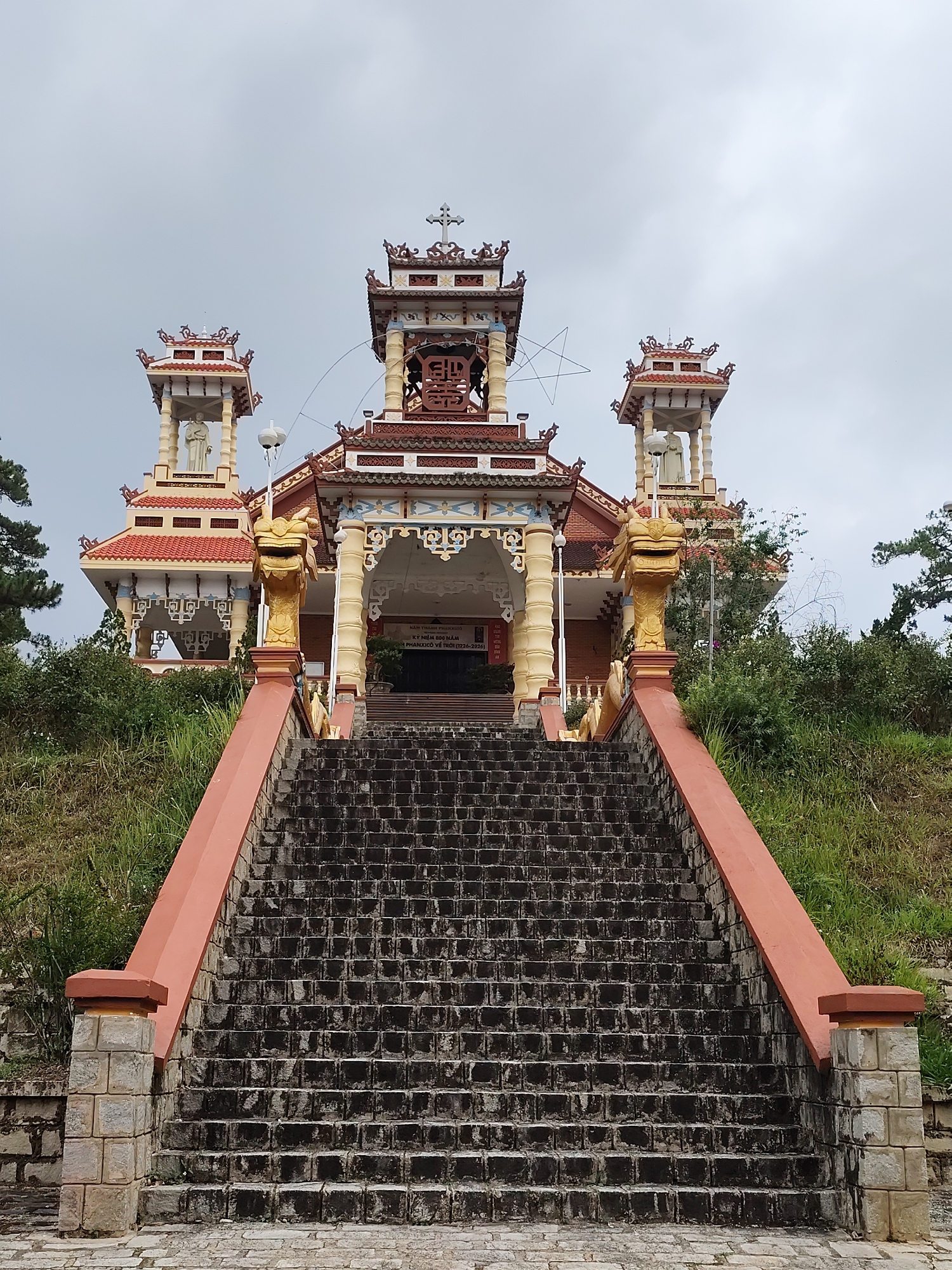 A wide shot depicts a temple in Xuân Hương - Đà Lạt, Vietnam. A long, wide set of dark grey stairs leads up a grassy hill to a three-tiered temple with intricate architectural details. The stairs are flanked by red railings with stone block pillars at the base. Golden dragon statues adorn the sides of the entrance. The temple has red-tiled roofs, ornate carvings, and religious symbols, including a cross at the top. The central tier has an opening leading to a shadowed interior, while the two side tiers feature statues under small pagoda-like roofs. The sky is overcast, and greenery surrounds the base of the temple.