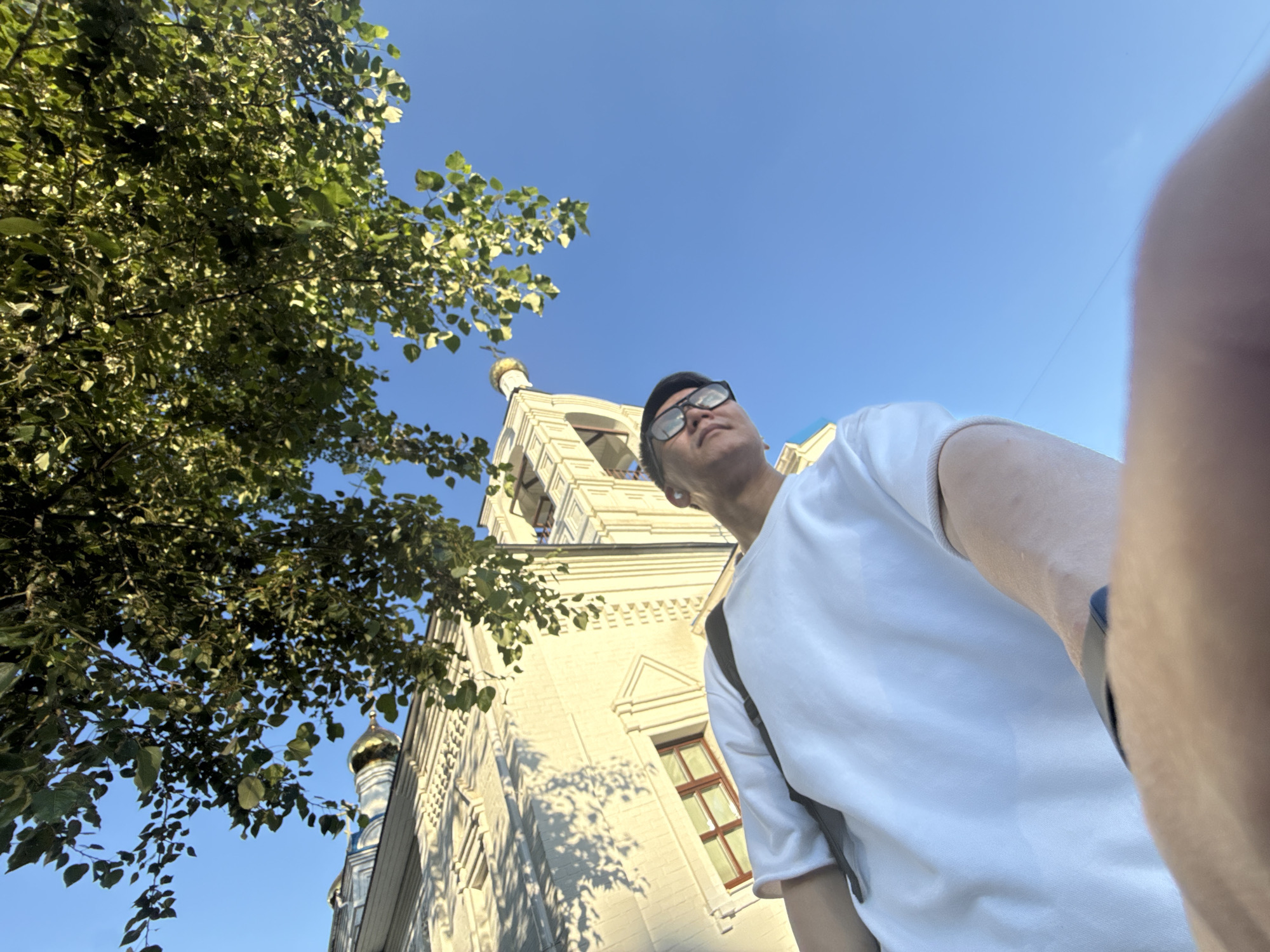 The media file shows a low-angle, wide-shot selfie taken outdoors on a sunny day. The primary subject is a person wearing a white t-shirt and glasses, with a dark baseball cap worn backwards. They are looking upwards and to the left, with their right arm extended, likely holding the camera. A portion of their arm and shoulder are visible in the foreground on the right side of the frame, and their torso is in the lower right half of the image.
In the background, a light-colored building with architectural details, possibly a church or historic structure, is visible. It has windows with brown frames and decorative elements along its facade. A golden dome with a cross on top is also visible on the building, suggesting a religious or historical site. To the left of the building, lush green tree branches with leaves are prominent, casting dappled shadows on the building. The sky is a clear, bright blue, indicating fair weather and daytime.
The perspective of the selfie, looking up at the subjects and the surroundings, creates a sense of looking out from a lower vantage point. The sunlight suggests it is likely daytime, possibly late morning or afternoon given the brightness of the sky and the shadows. The overall scene appears peaceful and captures a moment of observation or tourism. No text is visible in the image. The location context is Dolgoprudny, Russia.