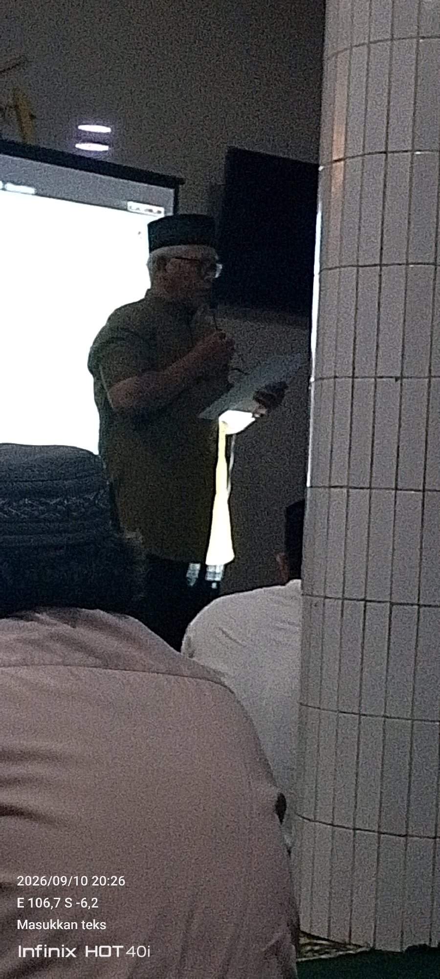 A man wearing a dark green shirt and a black peci hat stands in front of a projection screen. He is holding a microphone in his right hand and a document in his left. He appears to be speaking to an audience, some of whom are visible in the foreground. The setting appears to be an indoor event, possibly a lecture or presentation. The image also contains text overlay from a mobile phone, indicating the date as "2026/09/10 20:26" and coordinates "E 106,7 S -6,2". The text "Masukkan teks" and "Infinix HOT 40i" are also present. The coordinates suggest a location in Tangerang, Indonesia.