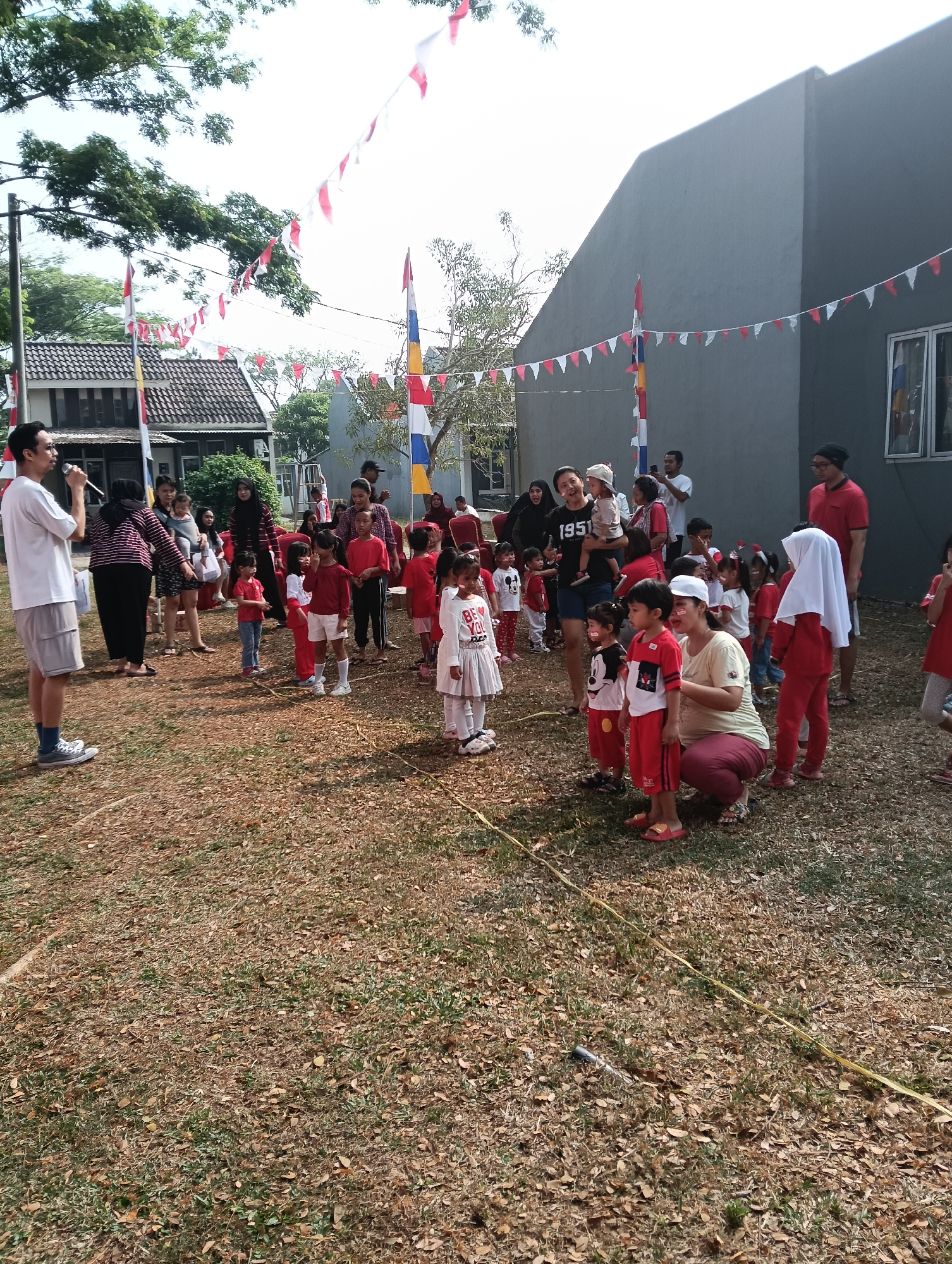 In Parung Panjang, Indonesia, a group of adults and children are gathered outdoors on a dry, grassy field adorned with red and white triangular flags strung overhead, likely for a national celebration. Several children, dressed in red and white outfits, are standing in a line, some with their faces painted. Adults, including a man holding a microphone, appear to be overseeing an event or activity. The background shows buildings, including a modern grey structure and a house with a tiled roof, and trees under a bright sky.
