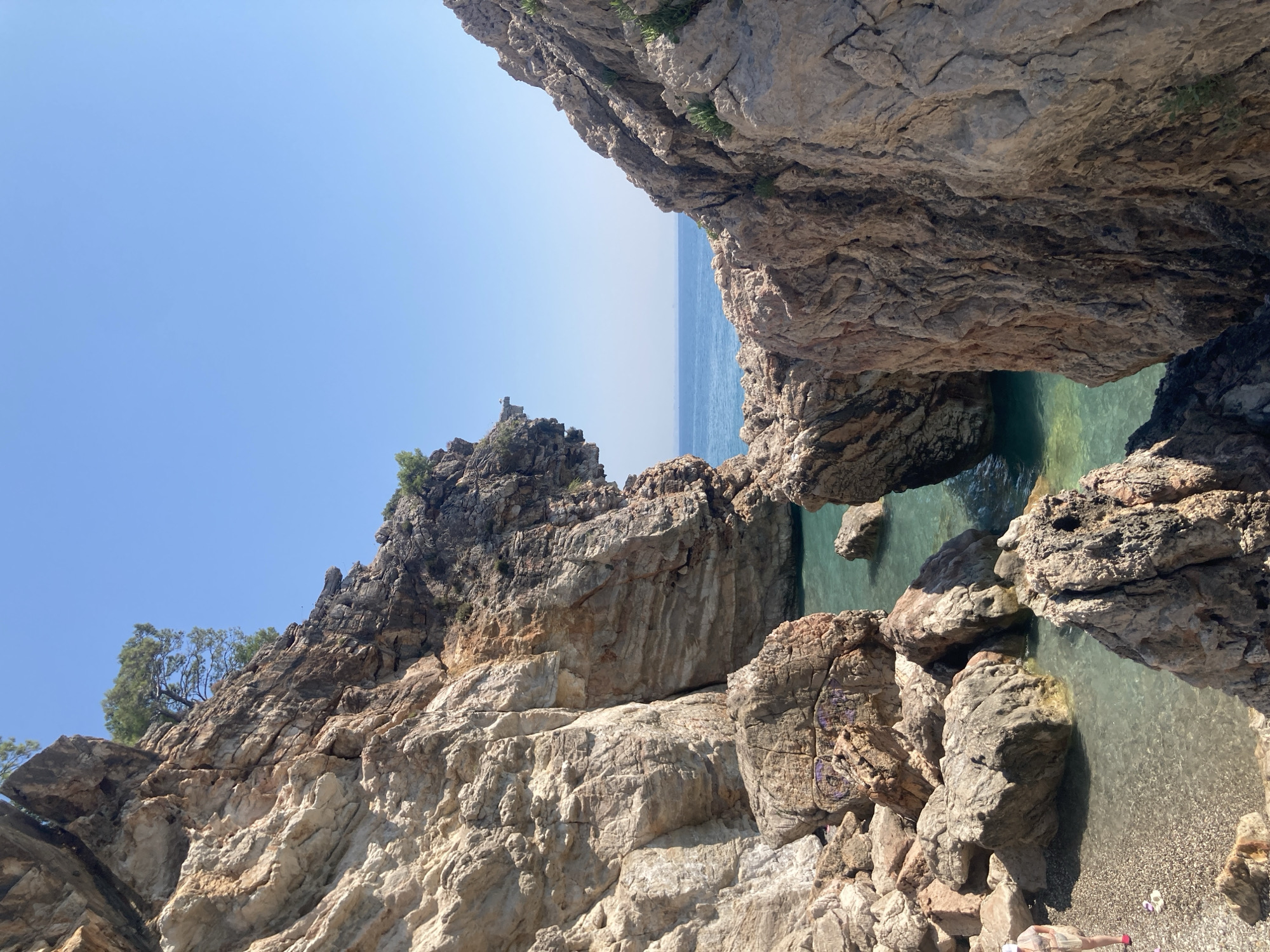 The image captures a scenic view of a secluded cove within the rocky coastline of Kemer, Turkey. The foreground is dominated by large, weathered rock formations of varying shades of brown and grey, some with visible striations and erosion. A small, pebble-strewn beach is visible on the left, where a single person wearing a light-colored hat and shorts stands near the water's edge. The water is a clear, turquoise hue, inviting and calm, with submerged rocks visible beneath the surface.
Beyond the cove, the sea stretches out to the horizon, meeting a clear, light blue sky. The imposing cliffs frame the view, with a sparse scattering of green vegetation clinging to their surfaces, indicating a rugged, natural environment. The overall impression is one of natural beauty and tranquility, typical of the coastal landscapes found in the Kemer region. The rock formations suggest geological activity and the passage of time, contributing to the raw, untamed character of the scene. The presence of the single individual emphasizes the peaceful and uncrowded nature of this particular spot.