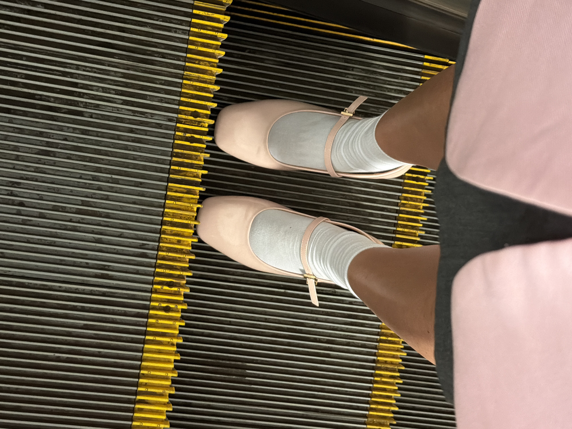 The image depicts a downward-facing view of a person standing on an escalator. The person is wearing pink, buckled ballet flats over white socks. The escalator has a dark gray surface with yellow side markers. The person's clothing is a combination of pink and gray. The location is within a city, but the specific place is not identifiable.