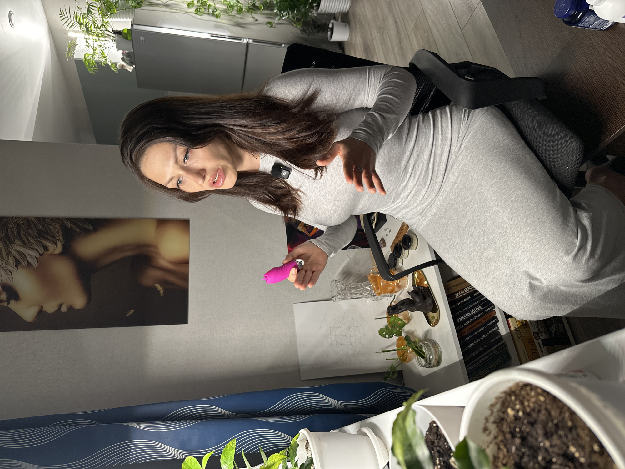 Here is a forensic description of the image:
A woman with long, dark hair and wearing a light gray, long-sleeved dress is seated in a chair. She appears to be holding a pink object in her left hand, possibly a personal massager or similar device. She is looking towards the object with a slight expression of interest or curiosity. A small microphone is clipped to her dress near her collarbone.
The setting seems to be a home or apartment interior. A piece of modern art is hanging on the wall behind her, featuring an abstract depiction of a face. A desk or shelf is visible to the right of the woman, with several items on top, including potted plants, books, and some decorative objects, arranged haphazardly. The desk appears to be cluttered.
Other elements in the background include a refrigerator and more plants. The overall lighting is somewhat dim. The flooring is a dark wooden finish.