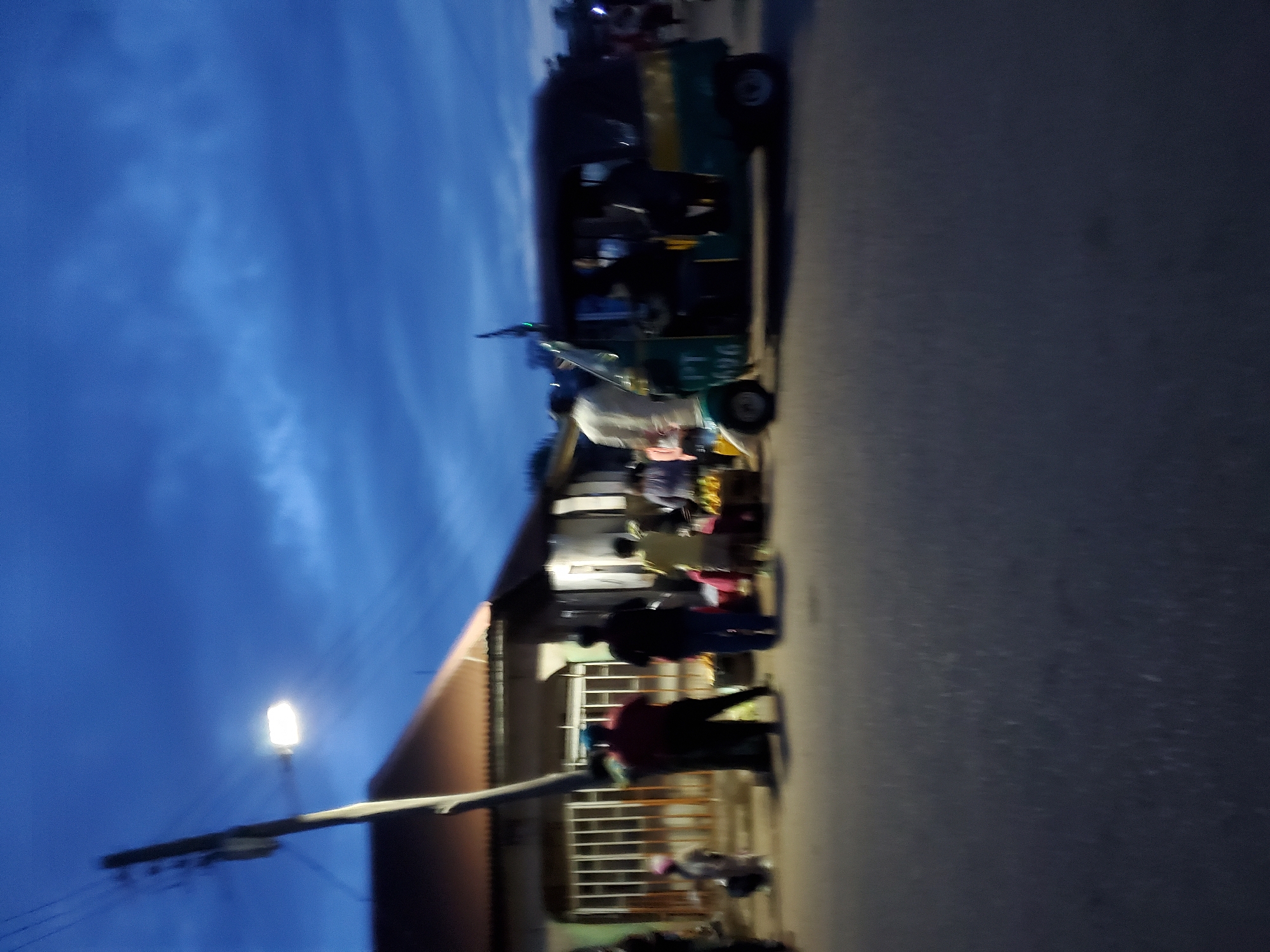 The image shows a street scene in Jos, Nigeria at dusk. The sky is blue with scattered clouds. A streetlight illuminates a building with a brown roof and a white barred window. Several people are standing and interacting near the building. A three-wheeled vehicle is parked on the street.