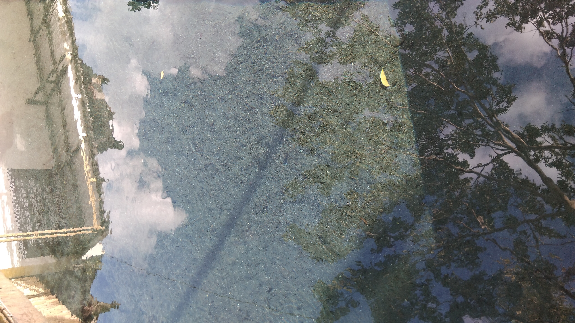The image depicts a shallow body of water, possibly a pond or a very still stream, reflecting the sky, clouds, and surrounding trees. The water's clarity allows a view of the pebbly bottom. The reflection shows a building with distinct architectural features and what appear to be yellow vertical poles. The overall impression is one of tranquility, with the reflections creating an abstract, layered visual. The location is identified as Ngaglik, Indonesia, suggesting an outdoor setting within that region.