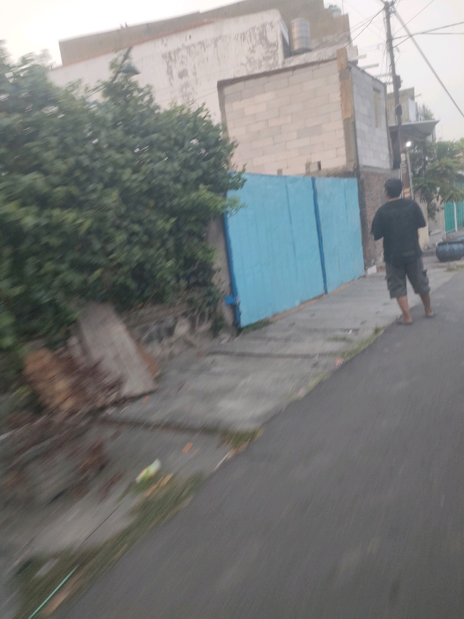 The image depicts a street scene in Surabaya, Indonesia, characterized by its urban environment. In the foreground, a person wearing a black t-shirt and patterned shorts walks away from the camera on an asphalt road. To the left of the road, a sidewalk is visible, bordered by a vibrant blue fence or wall. Behind the fence, a dense green bush or tree obscures much of the background. Further back, a partially constructed or aged building is visible, with visible concrete blocks and some discolored plaster. The sky above is overcast, suggesting a cloudy day. The image quality is somewhat blurred, possibly due to motion, but the elements of the scene are discernible.