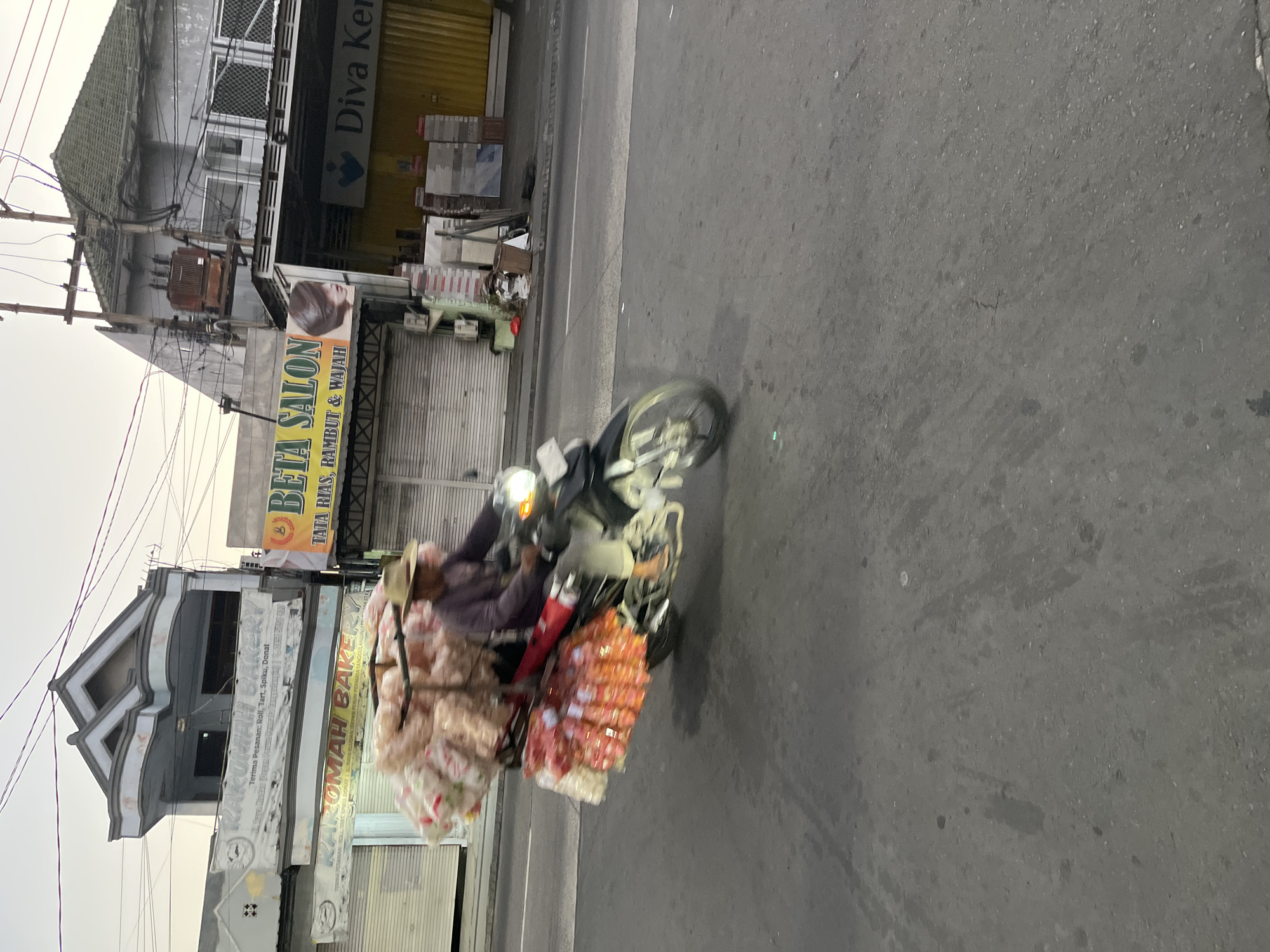 In Boro, Indonesia, a man wearing a hat rides a motorcycle laden with numerous bags of snacks. The motorcycle is captured in motion, with the wheels blurred due to the speed. The man is dressed in a long-sleeved shirt and pants. The background features buildings with signage, including "KAROMAH BAKERY" and "BETA SALON." The sky is overcast, and the street appears wet in places, suggesting recent rain or damp conditions. Electrical wires are visible crisscrossing above the street.