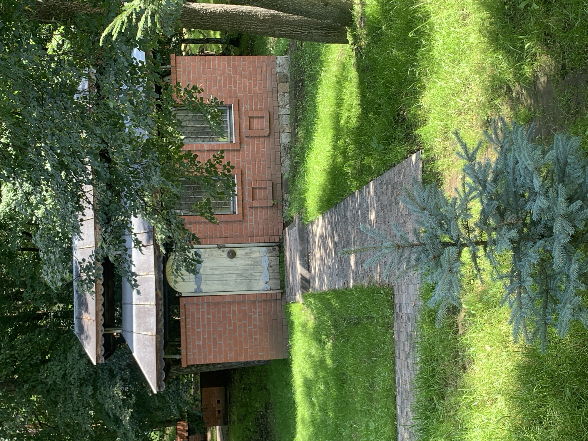 The image depicts a scenic outdoor setting in Arbuzov-Baran, Russia. The main subject is a small, rustic brick building with a light-colored wooden door and barred windows. Adjacent to the building, a pathway made of paving stones leads through a lush green lawn. A young blue spruce tree stands on the right side of the pathway, with its branches extending towards the camera. The background is dominated by dense green foliage from trees, suggesting a secluded, natural environment. The sunlight filtering through the trees casts dappled shadows on the grass and pathway, indicating it is likely daytime, possibly late morning or early afternoon. There are no people visible in the image. The overall atmosphere is tranquil and serene, characteristic of a garden or a rural backyard. The building appears to be a functional structure, possibly a shed or a small outbuilding, blending harmoniously with the natural surroundings. The paving stones and the brickwork suggest a well-maintained area, while the abundant greenery points to a temperate climate.